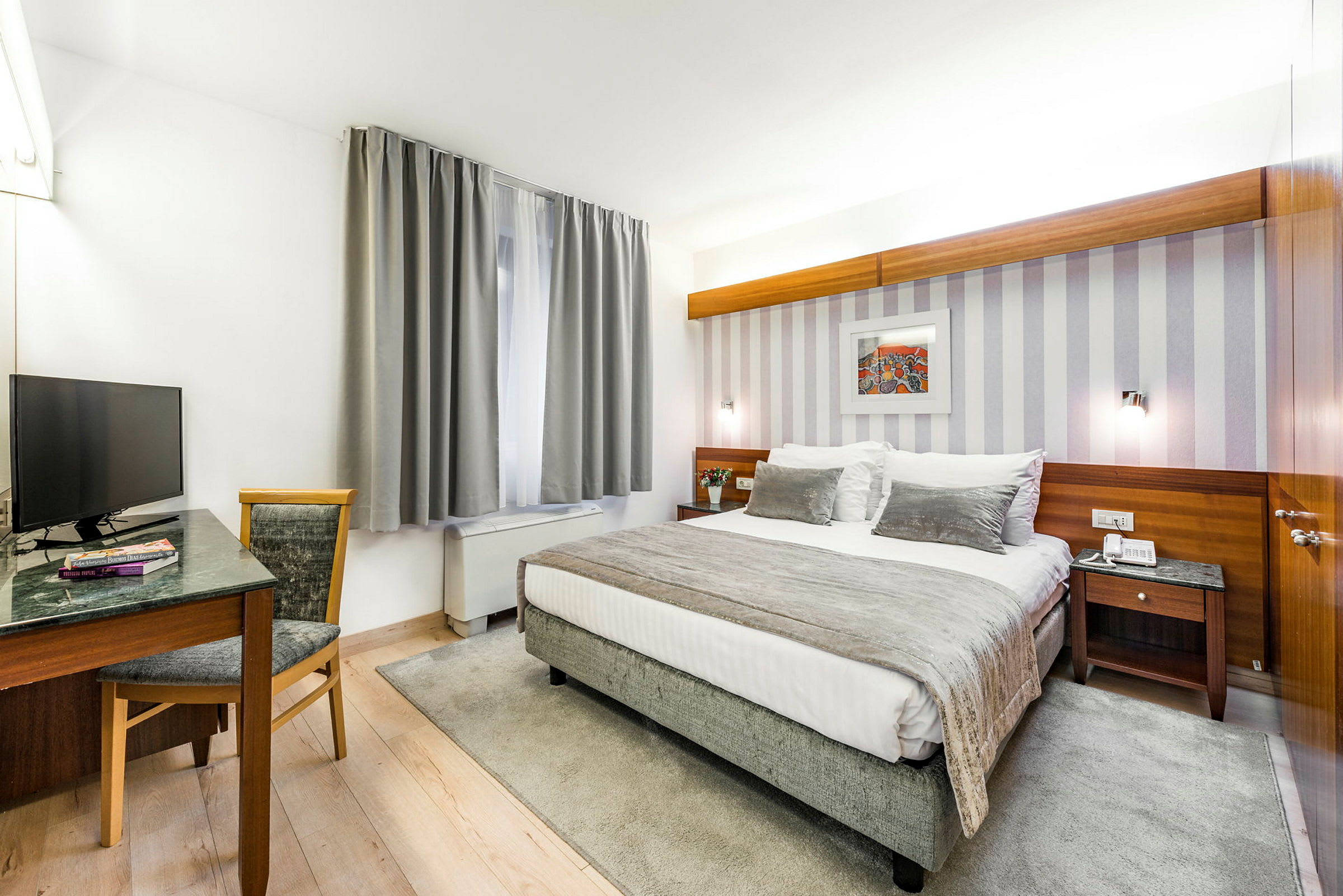 Et hotellrom med en dobbeltseng og et skrivebord ved siden av et vindu med grå gardiner. (AI-generert tekst)