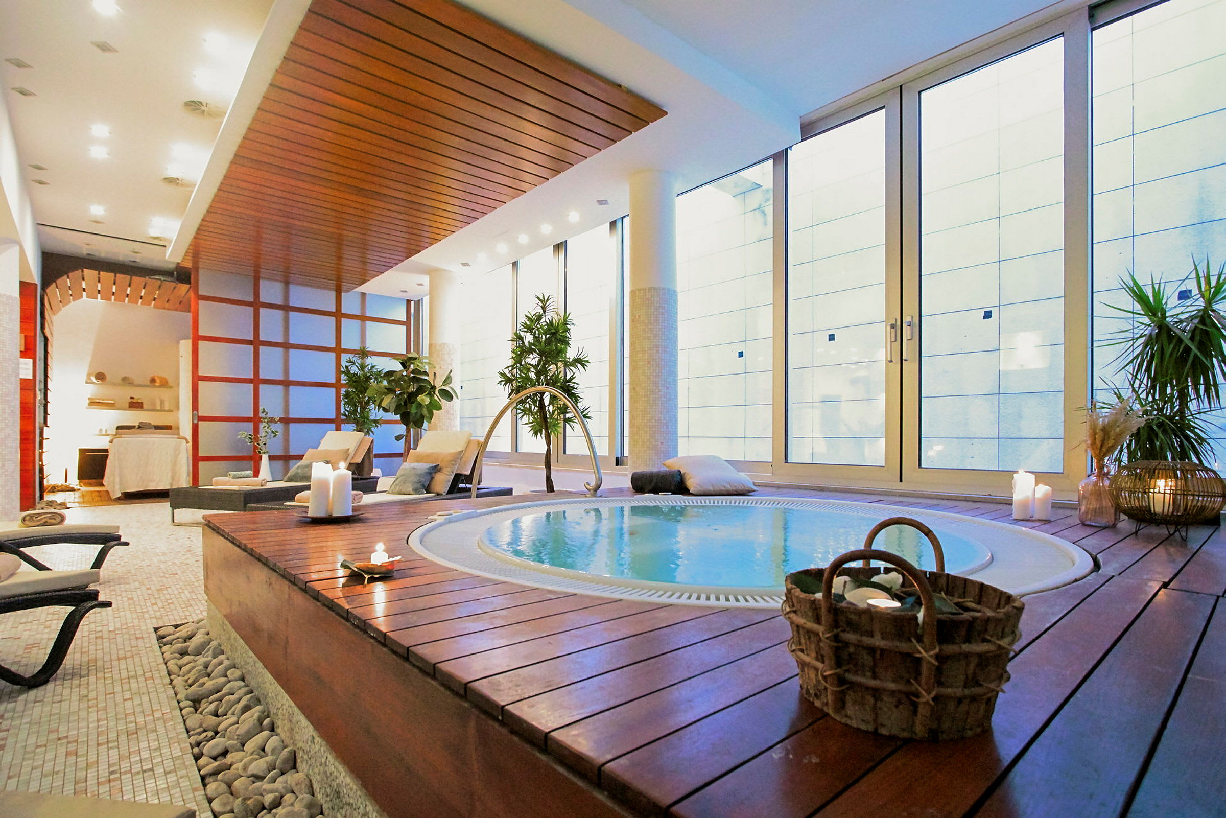 Et elegant spa med en jacuzzi omgitt av trepaneler, stoler og levende planter. Store glassvegger gir naturlig lys. (AI-generert tekst)