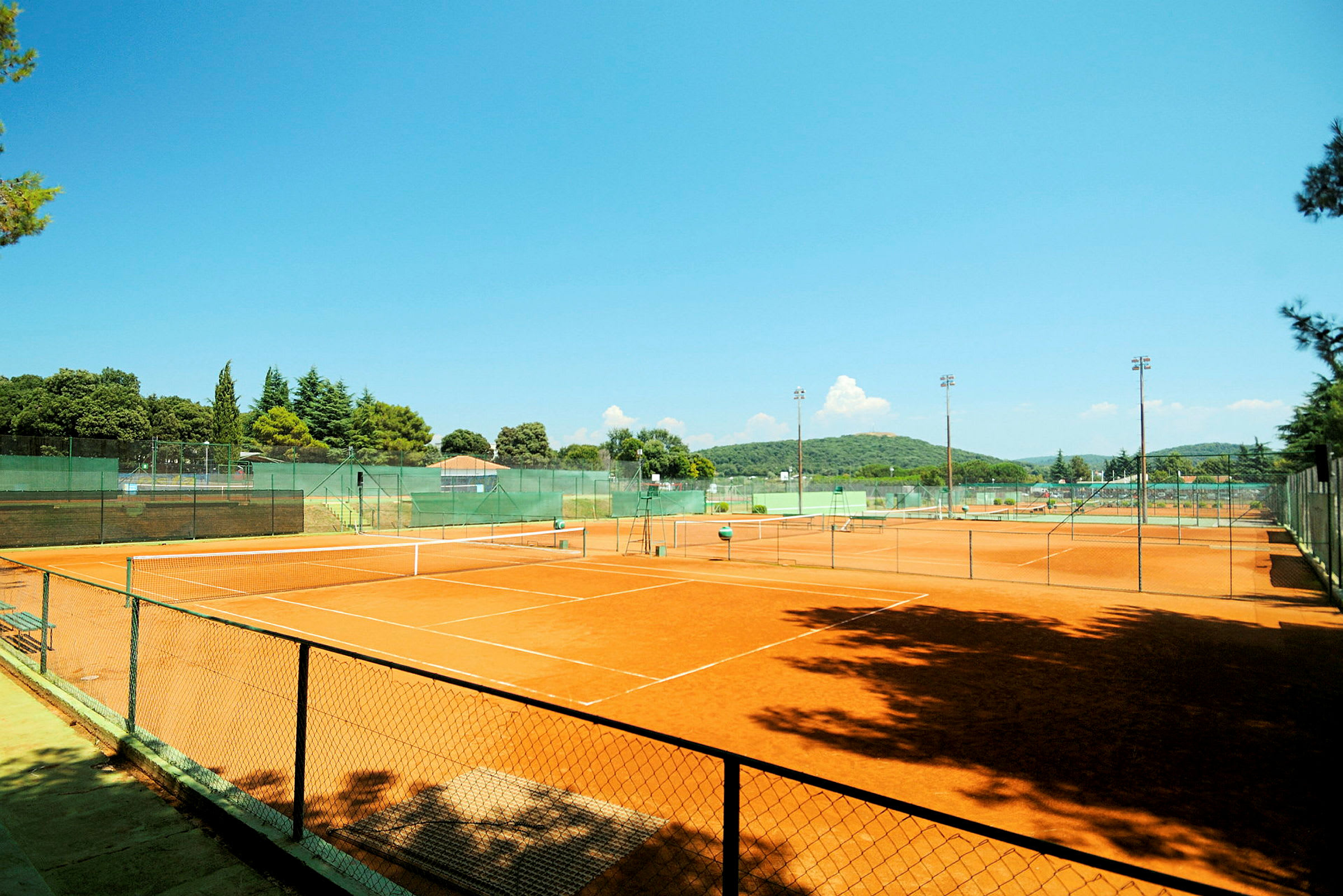Bilde av en utendørs tennisbane med grusunderlag, omgitt av grøntområder og et gjerde. (AI-generert tekst)