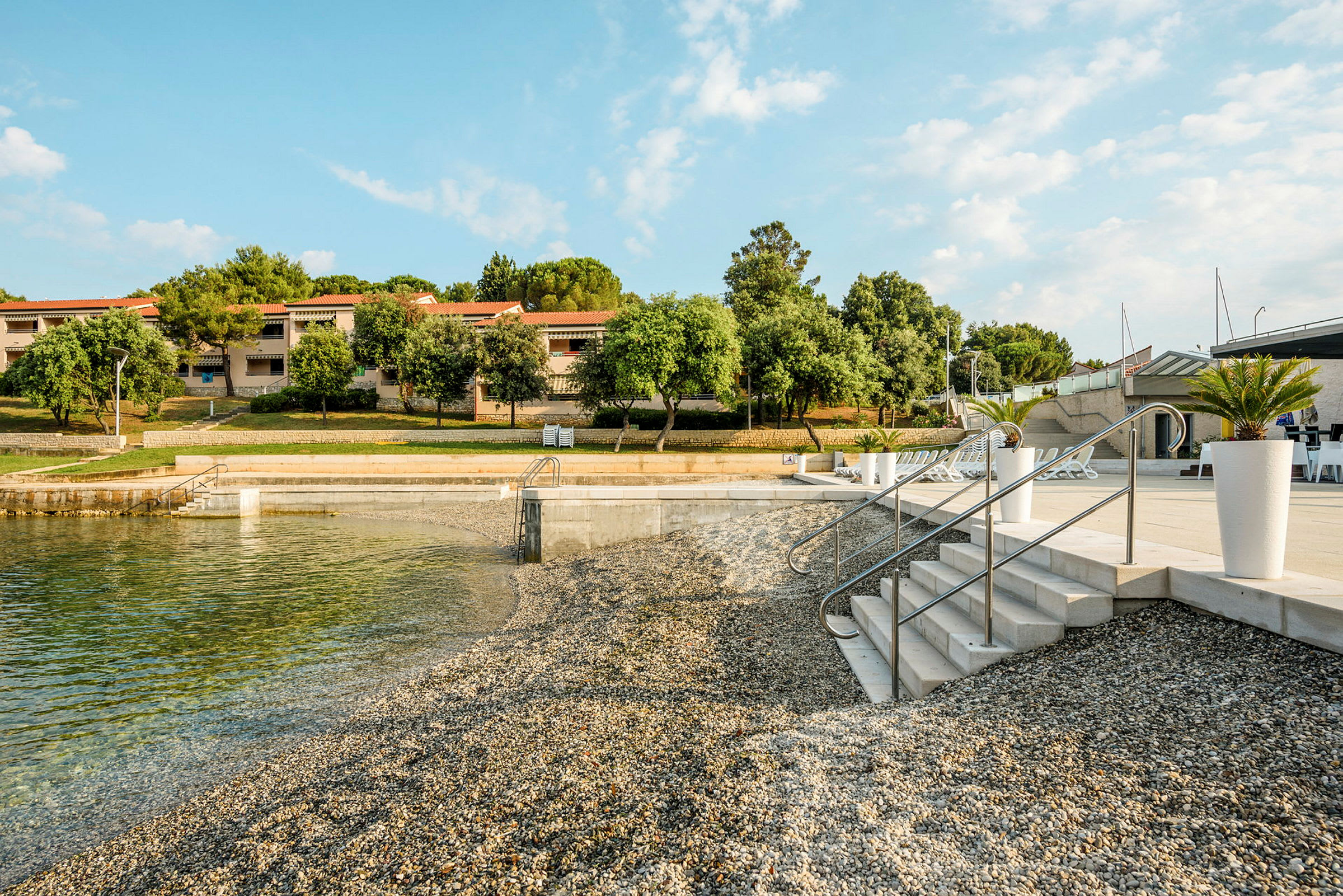 En strand med småstein og klart vann, med trappetrinn som fører ned til vannet. I bakgrunnen ses hus med røde tak og frodig vegetasjon. (AI-generert tekst)