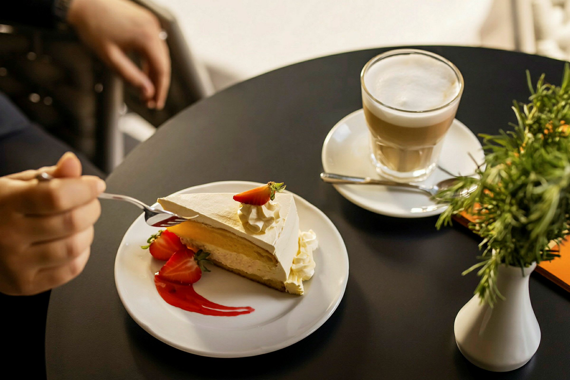 En desserttallerken med et stykke kake pyntet med jordbær og krem, ved siden av en kaffekopp på et svart bord. (AI-generert tekst)