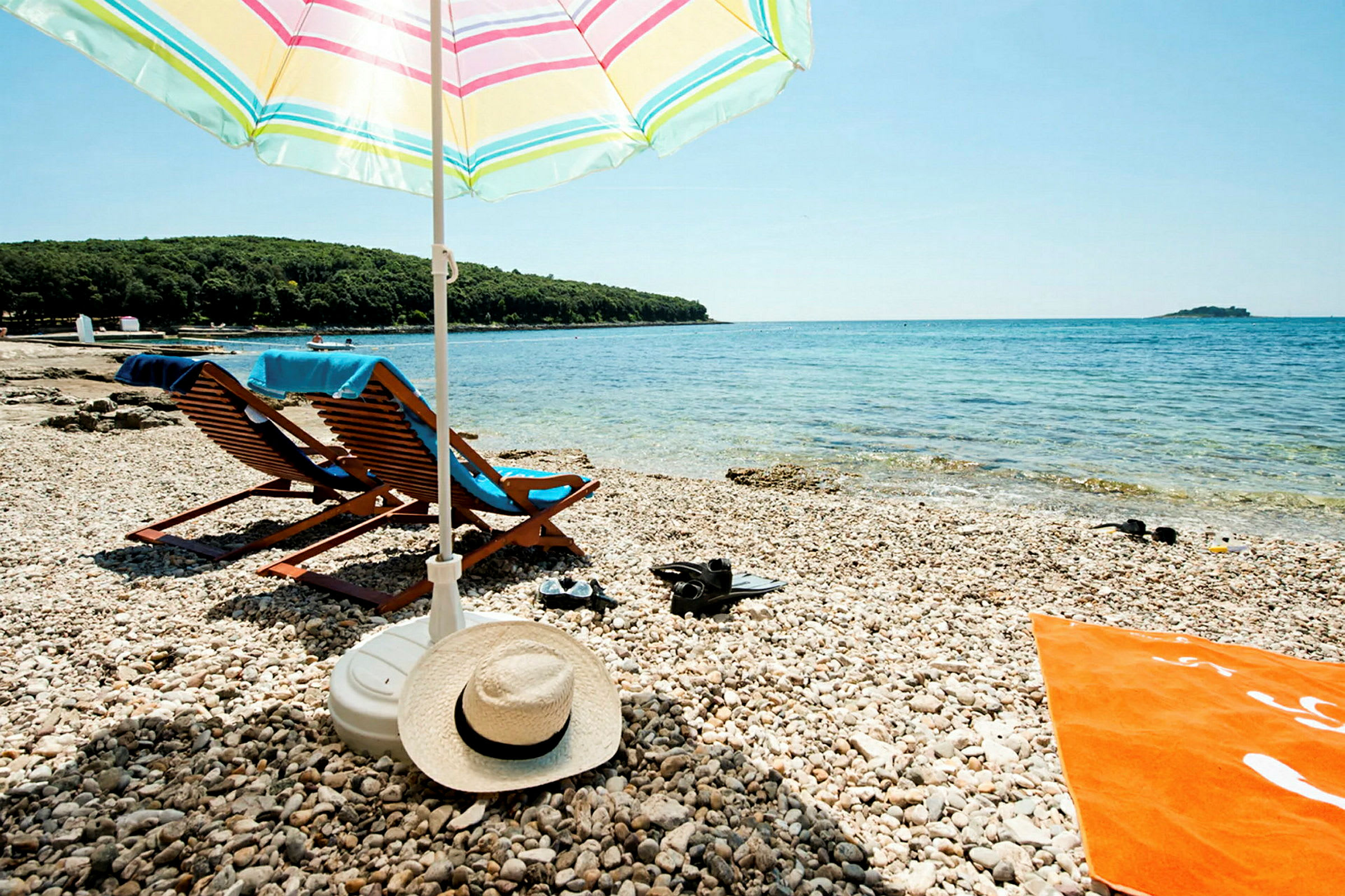 En steinstrand med to solsenger under en stripet parasoll. Det turkise havet kan sees i bakgrunnen. (AI-generert tekst)