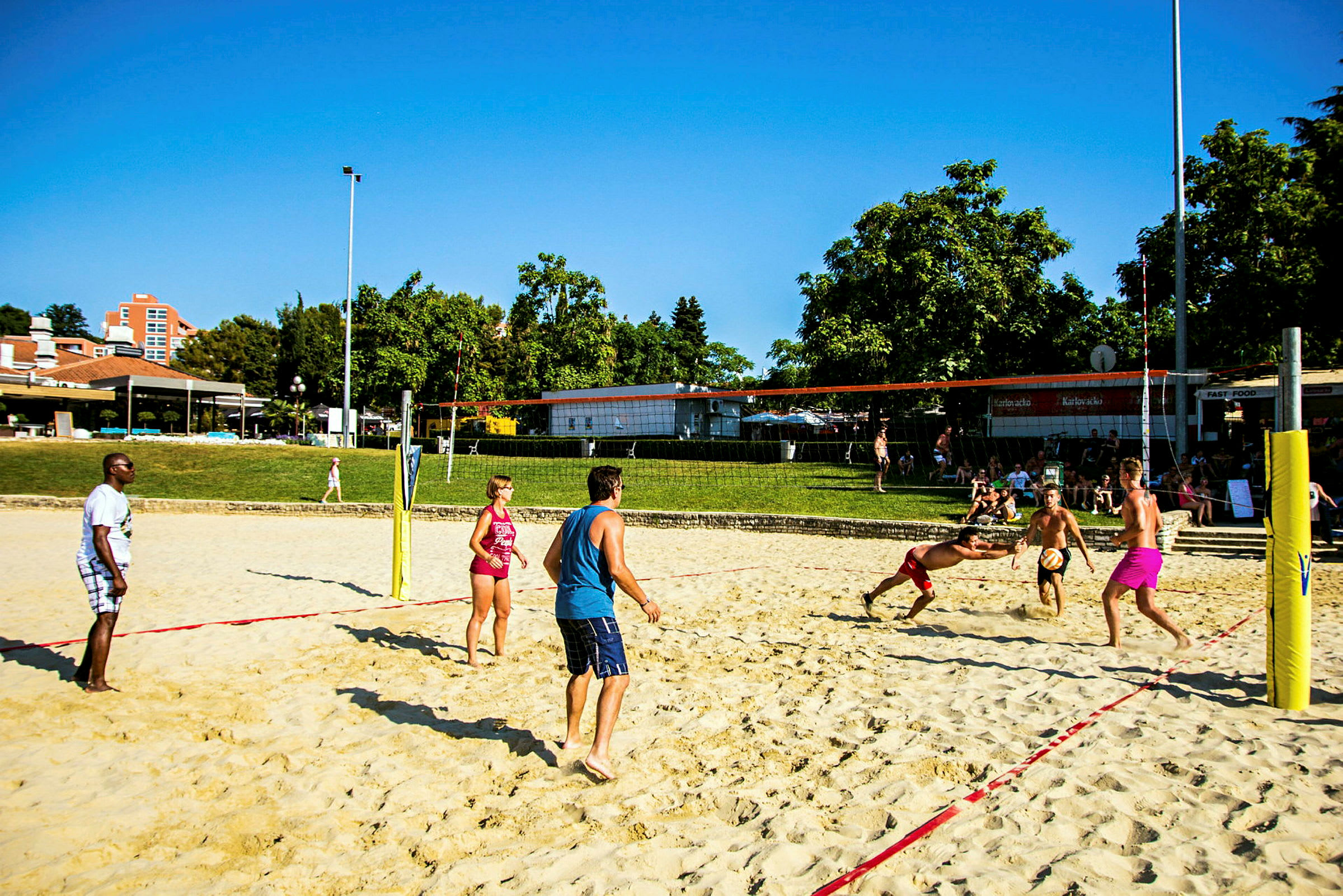 Folk spiller sandvolleyball på en sandbane i et utendørs område med trær og bygninger i bakgrunnen. (AI-generert tekst)