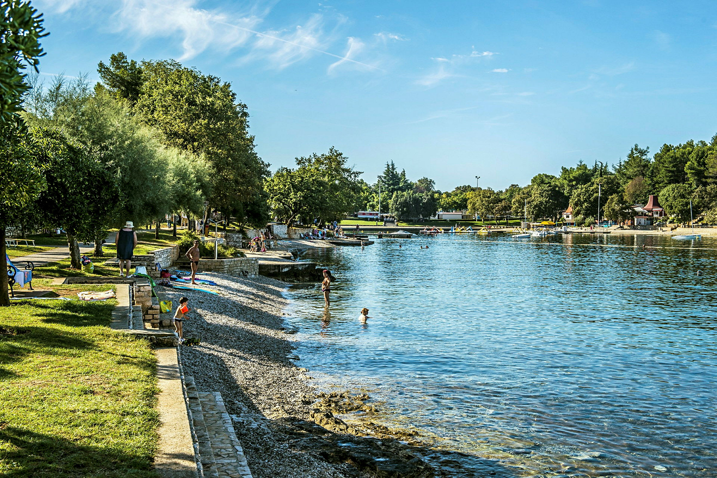 Bilde av en steinstrand ved en rolig bukt. Flere personer kan ses ved vannkanten og på gressplen ved siden av strandpromenaden. (AI-generert tekst)