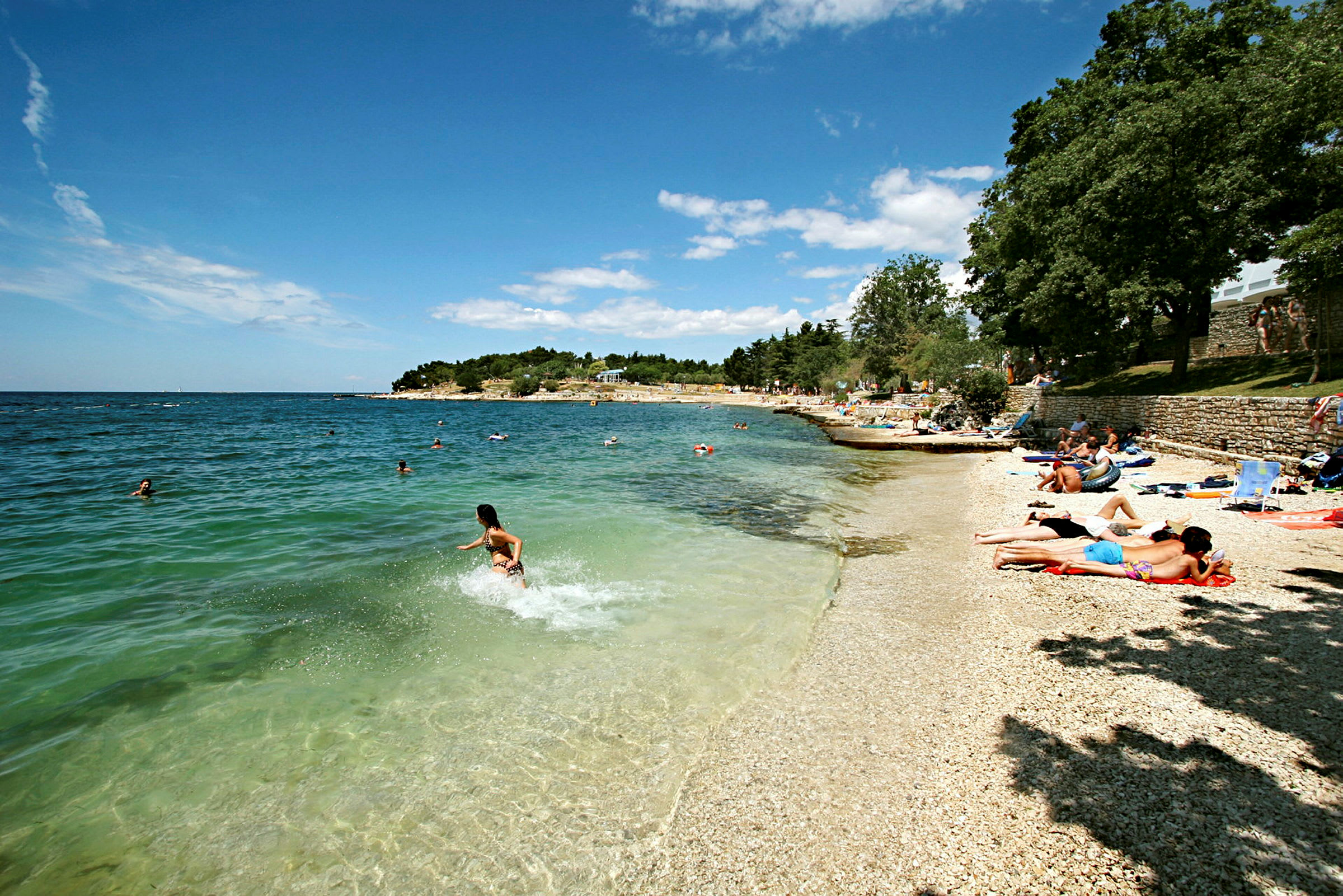 En strand med småstein hvor folk soler seg og bader i det klareblå havet. (AI-generert tekst)