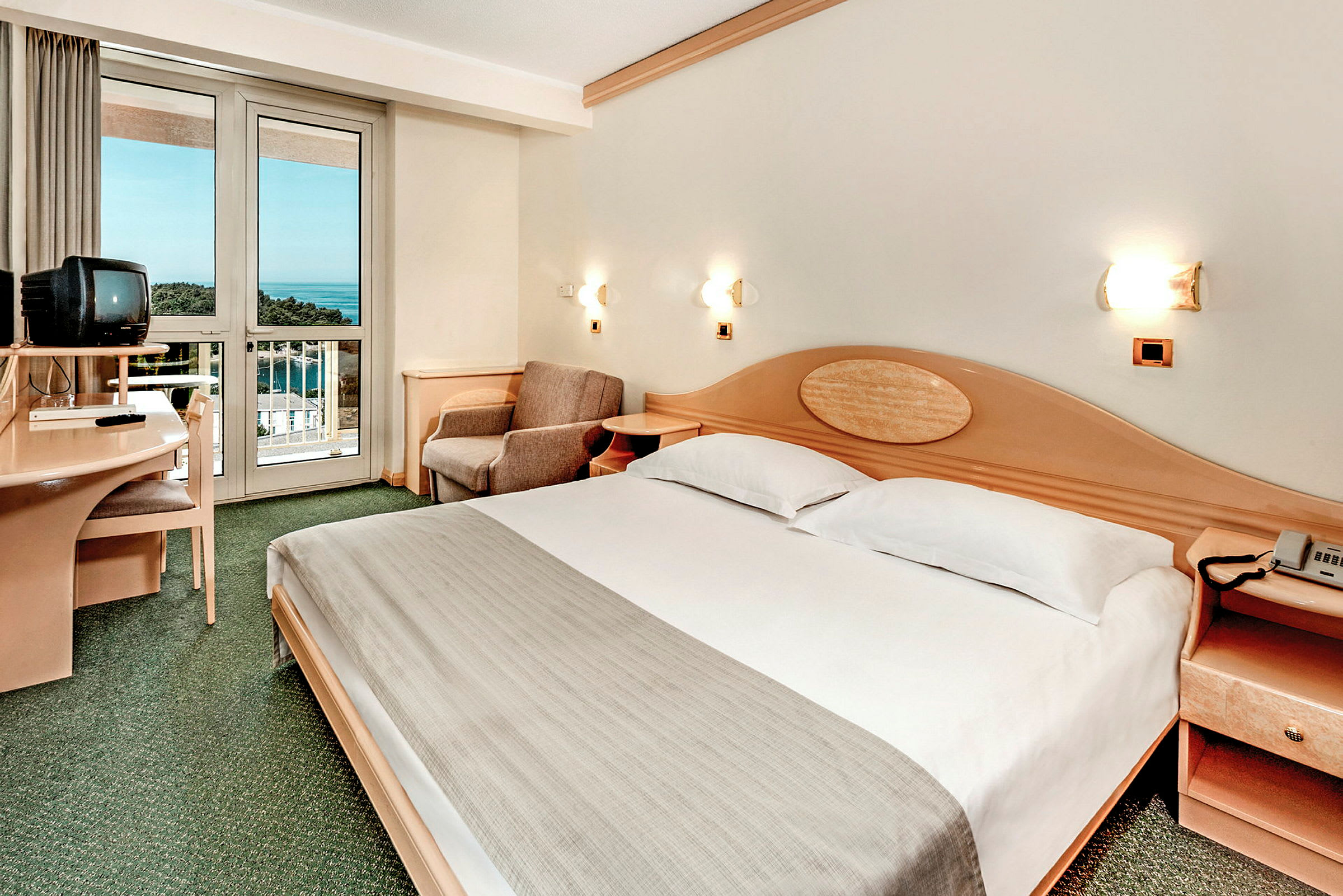 Et hotellrom med en dobbeltseng, en TV på et skrivebord og en balkong med utsikt over havet. (AI-generert tekst)