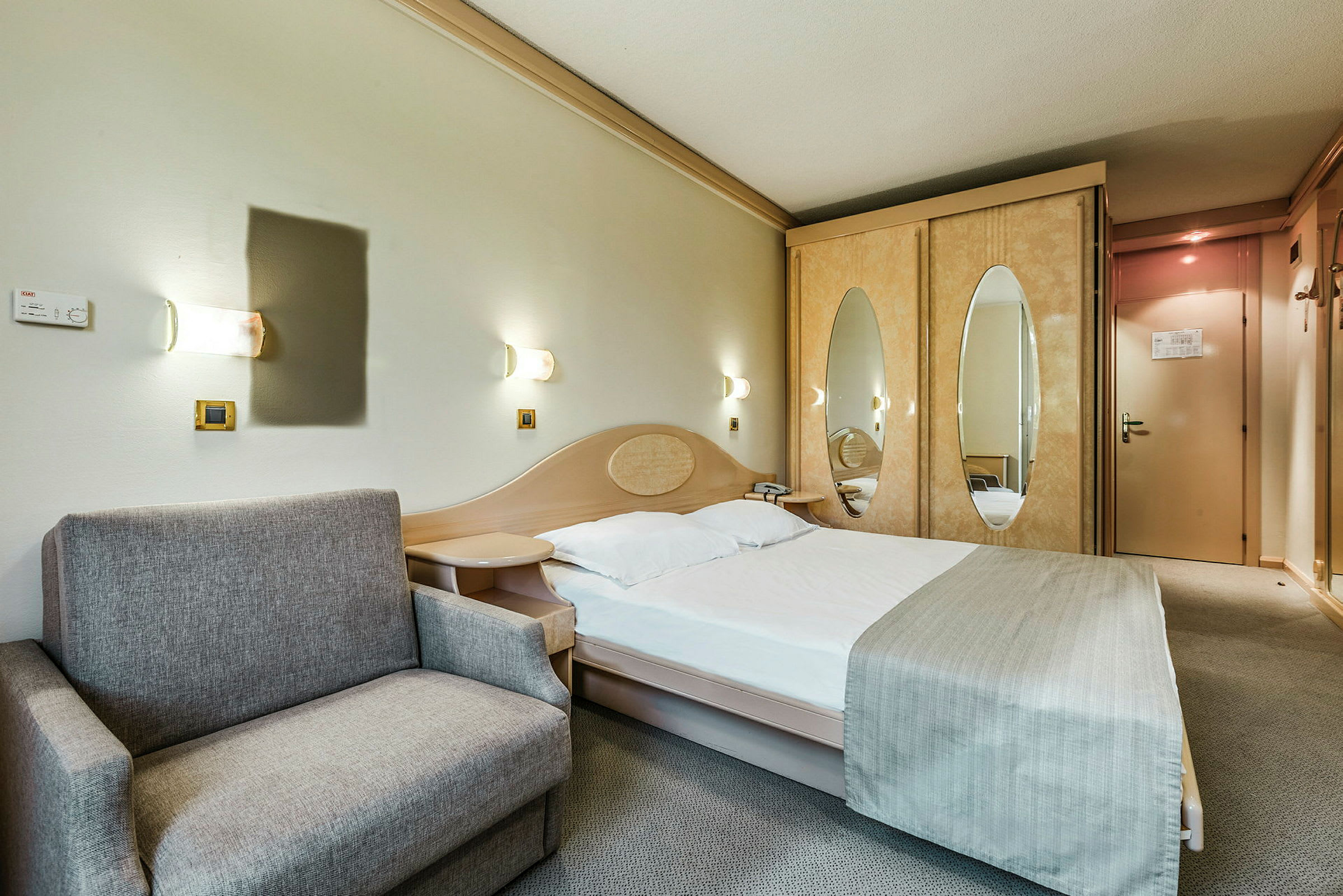 Et hotellrom med en oppredd seng, en grå sofa og et skap med speildører. (AI-generert tekst)