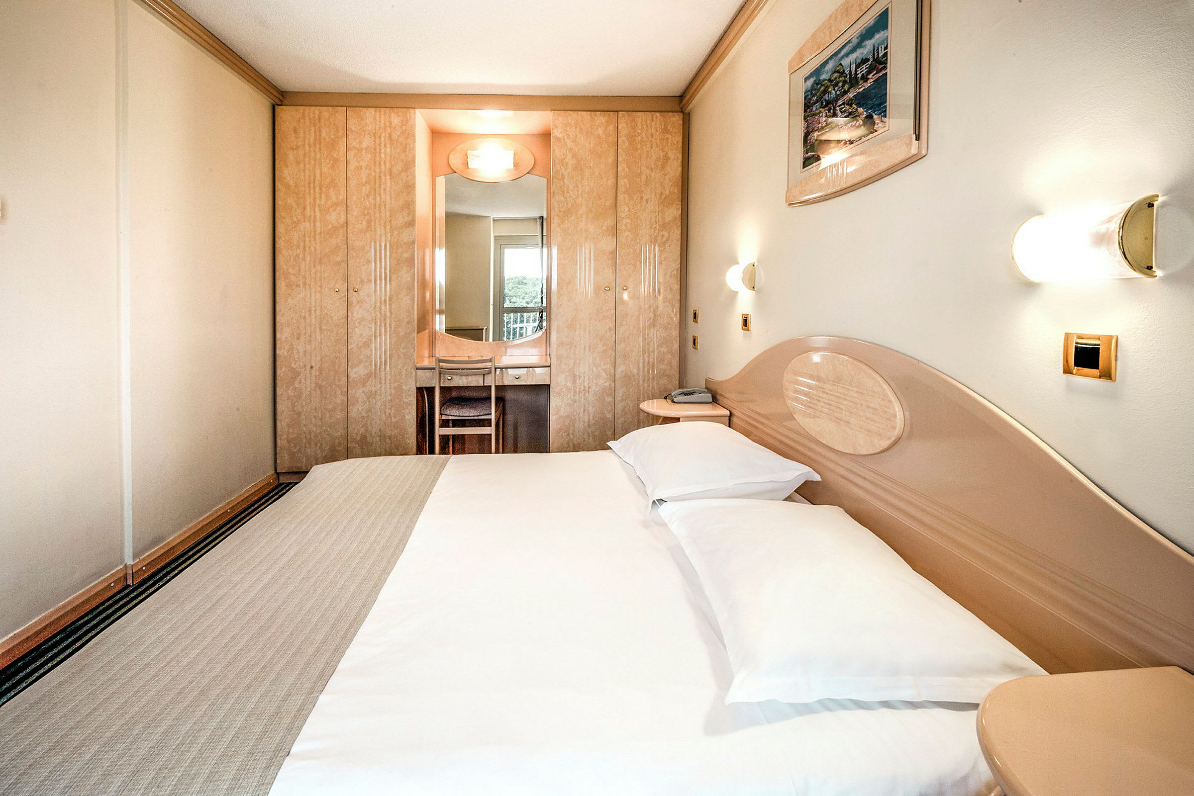 Et hotellrom med en dobbeltseng, veggmonterte lamper og et innebygd skap med et speil foran en stol. (AI-generert tekst)