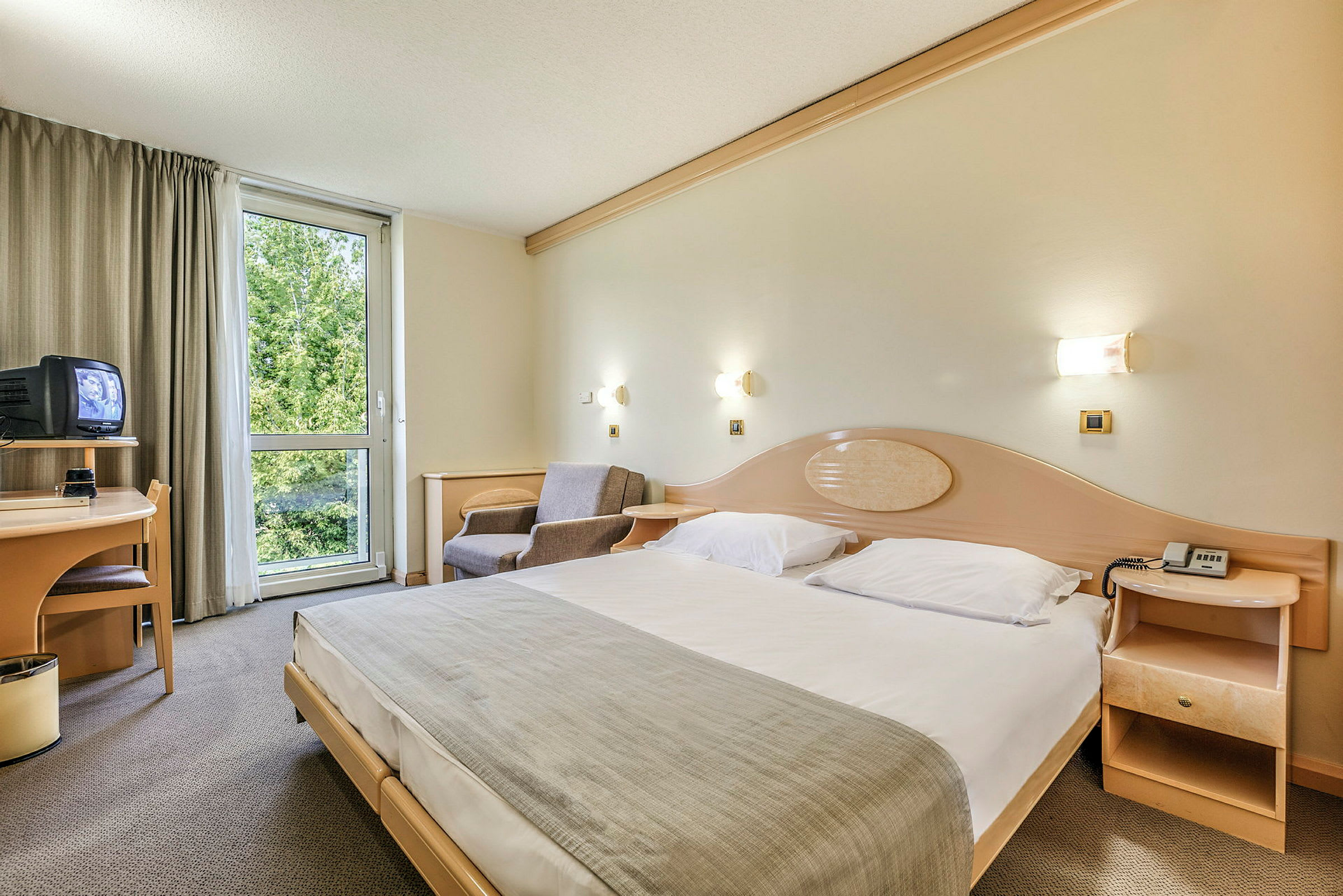 Hotellrom med en stor seng, en lenestol, et skrivebord med en TV og et vindu med utsikt mot grøntområder. (AI-generert tekst)