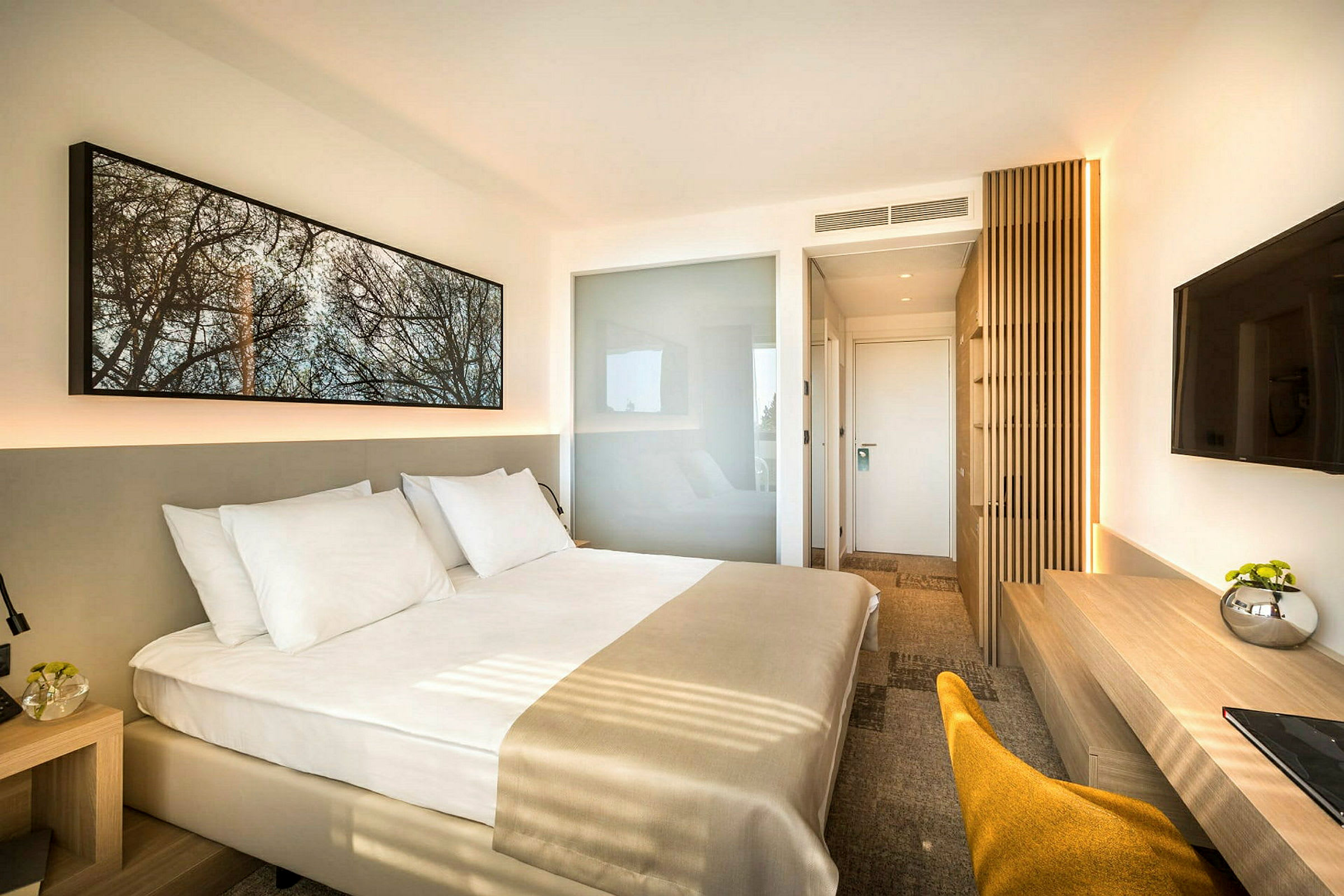 Et moderne hotellrom med en stor seng med hvitt sengetøy. Rommet har en minimalistisk innredning med en veggmontert TV, et skrivebord og en stol. (AI-generert tekst)