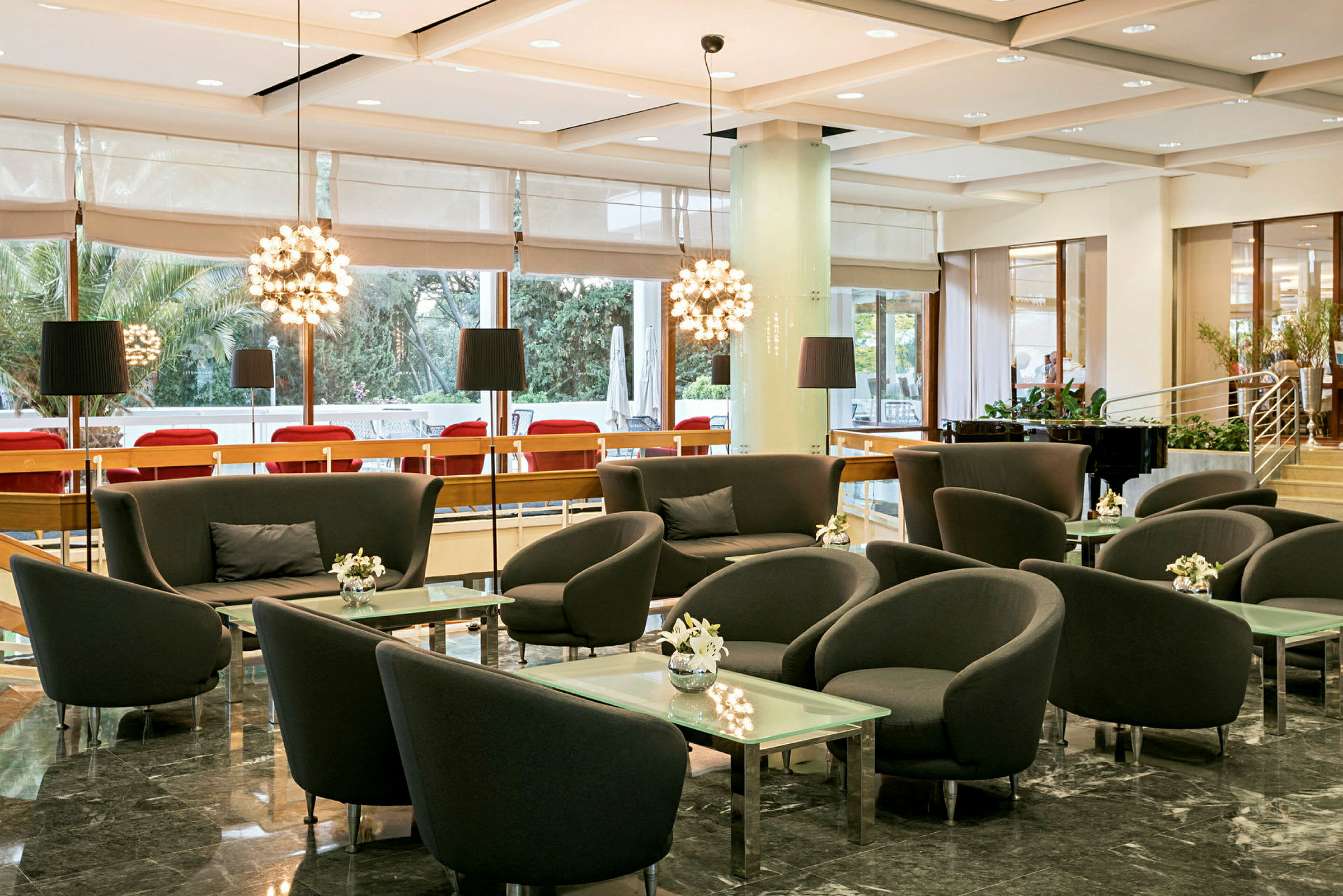 En godt opplyst lounge med sittegrupper bestående av lenestoler og sofaer rundt glassbord, omgitt av store vinduer og moderne interiør. (AI-generert tekst)