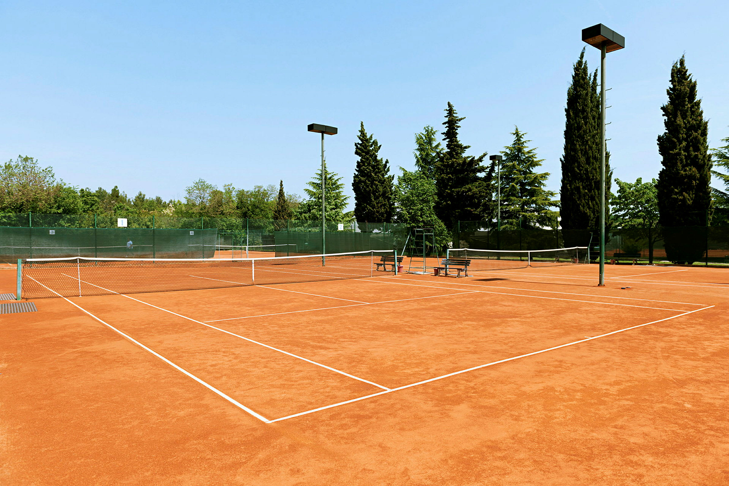 En tennisbane med grusunderlag omgitt av trær og gjerder. (AI-generert tekst)