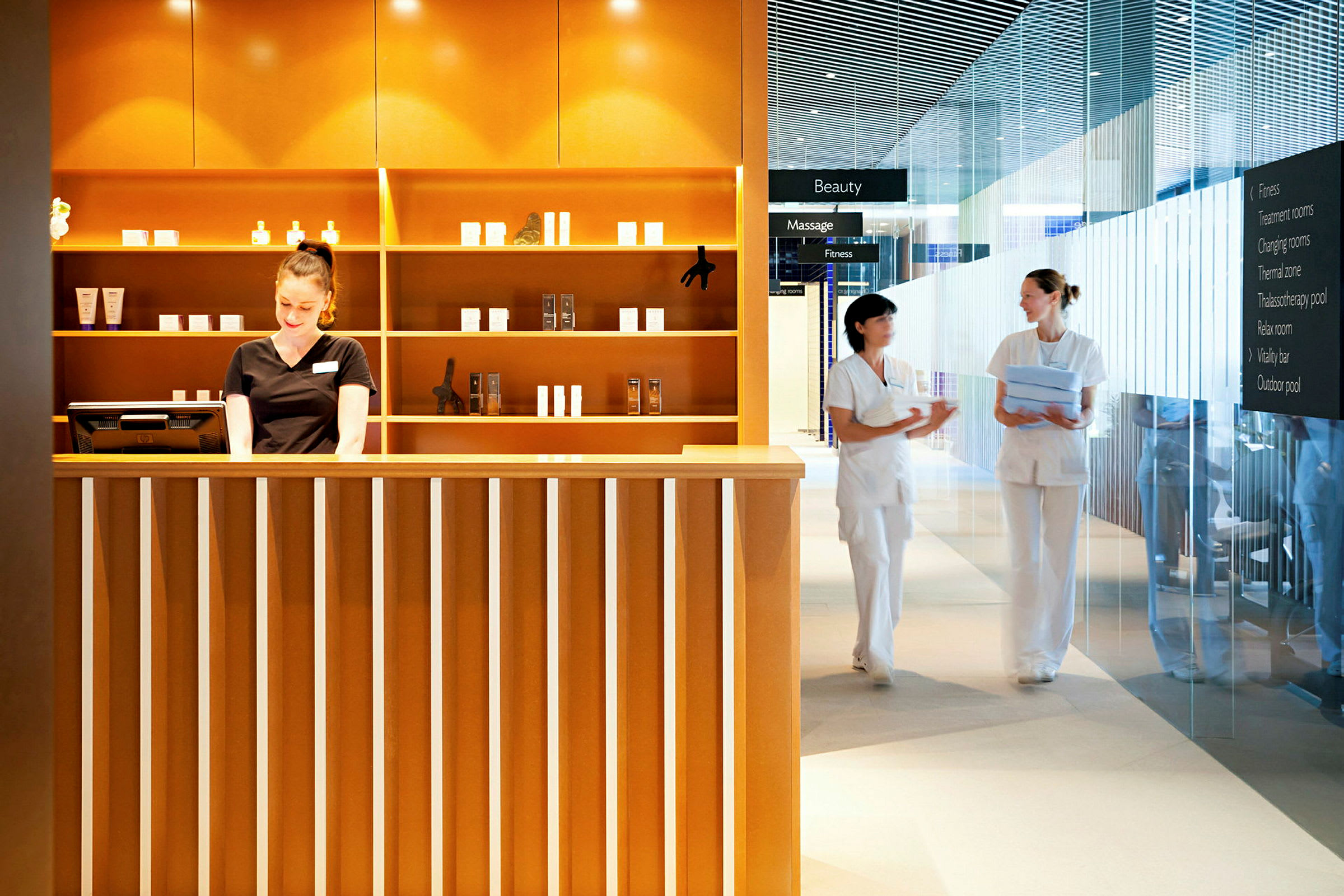 Resepsjon for spa- eller skjønnhetsanlegg med hyller fylt med produkter og to personer i arbeidsklær som samtaler ved siden av. (AI-generert tekst)