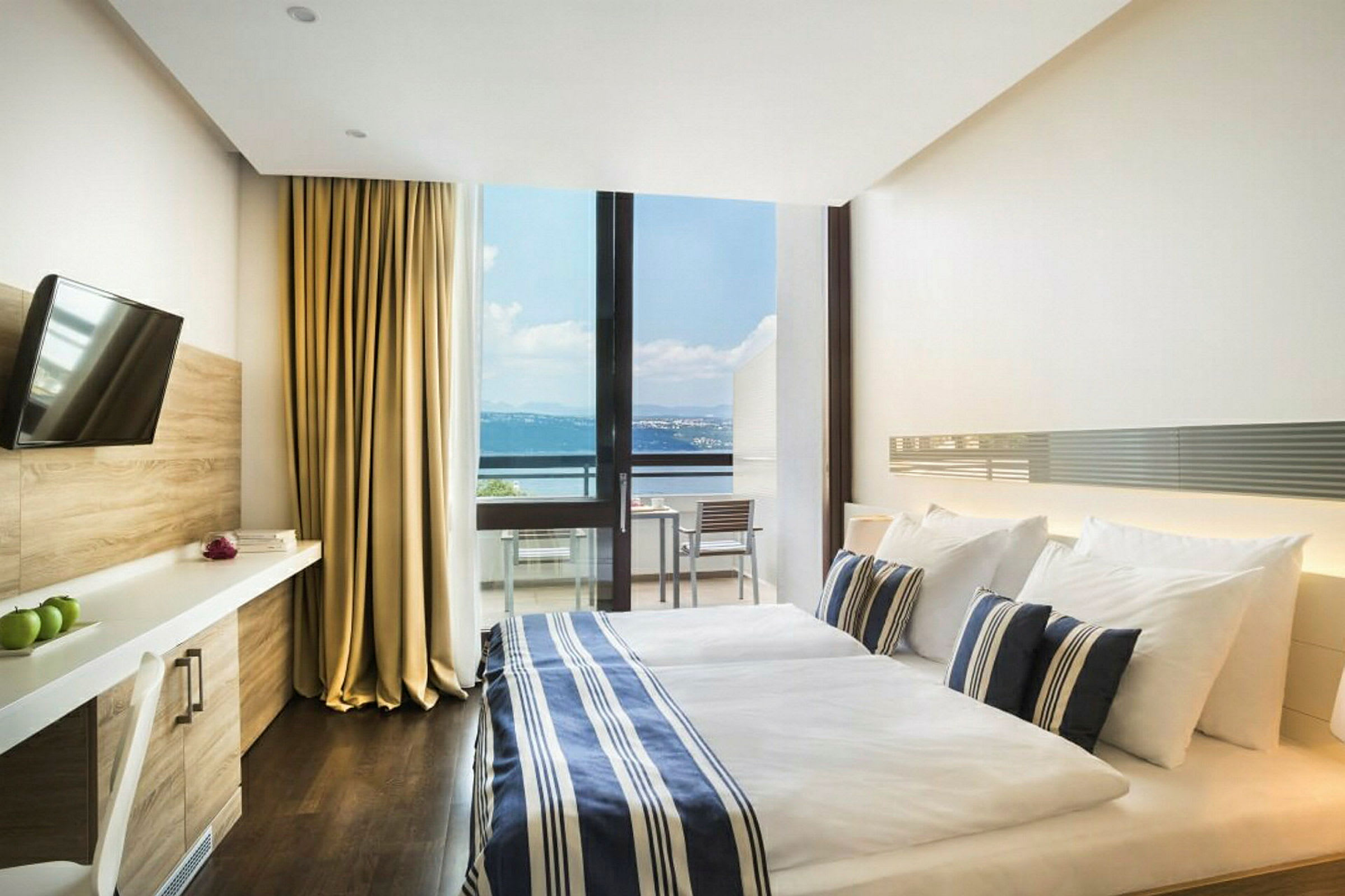 Et hotellrom med en dobbeltseng, sengetøy i blått og hvitt stripemønster, og en stor glassvegg som viser en balkong med utsikt over havet. Rommet har også en TV og skrivebord. (AI-generert tekst)