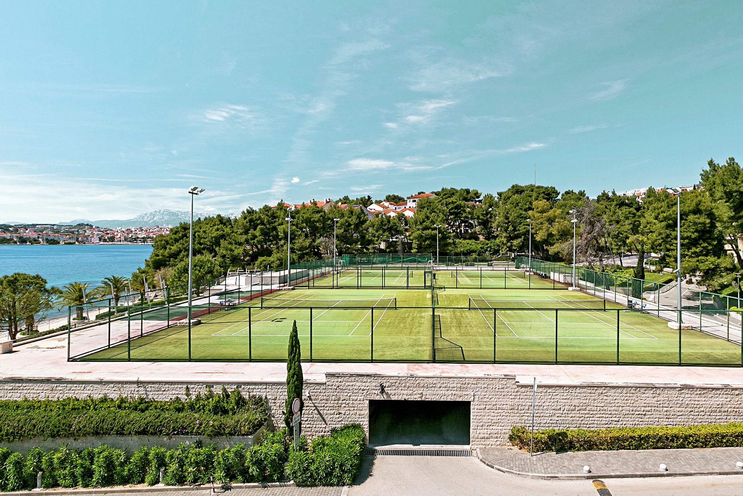 Bilde av utendørs tennisbaner med havet og bygninger i bakgrunnen. (AI-generert tekst)