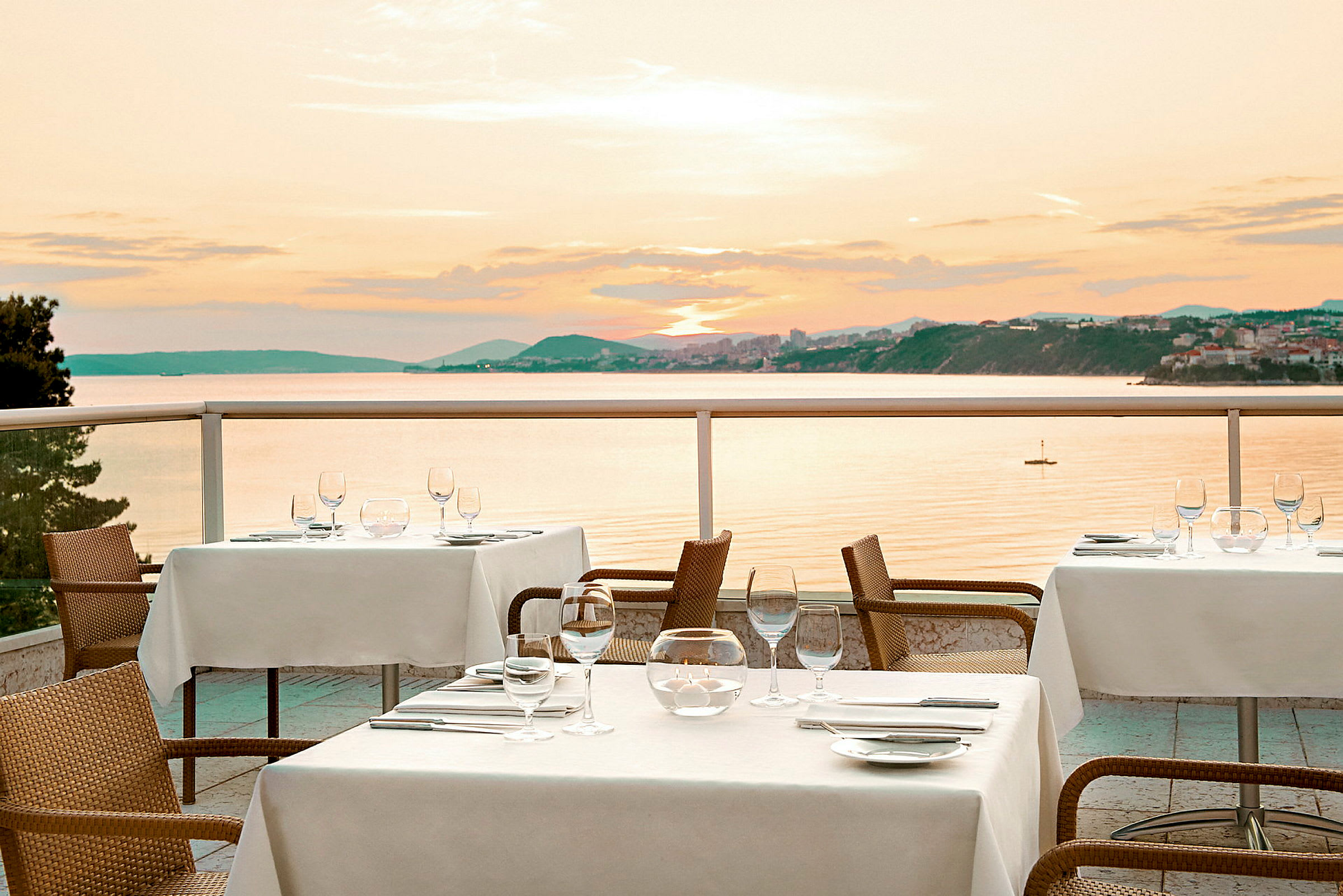 Bord med hvite duker på en terrasse med utsikt over havet og solnedgangen. (AI-generert tekst)