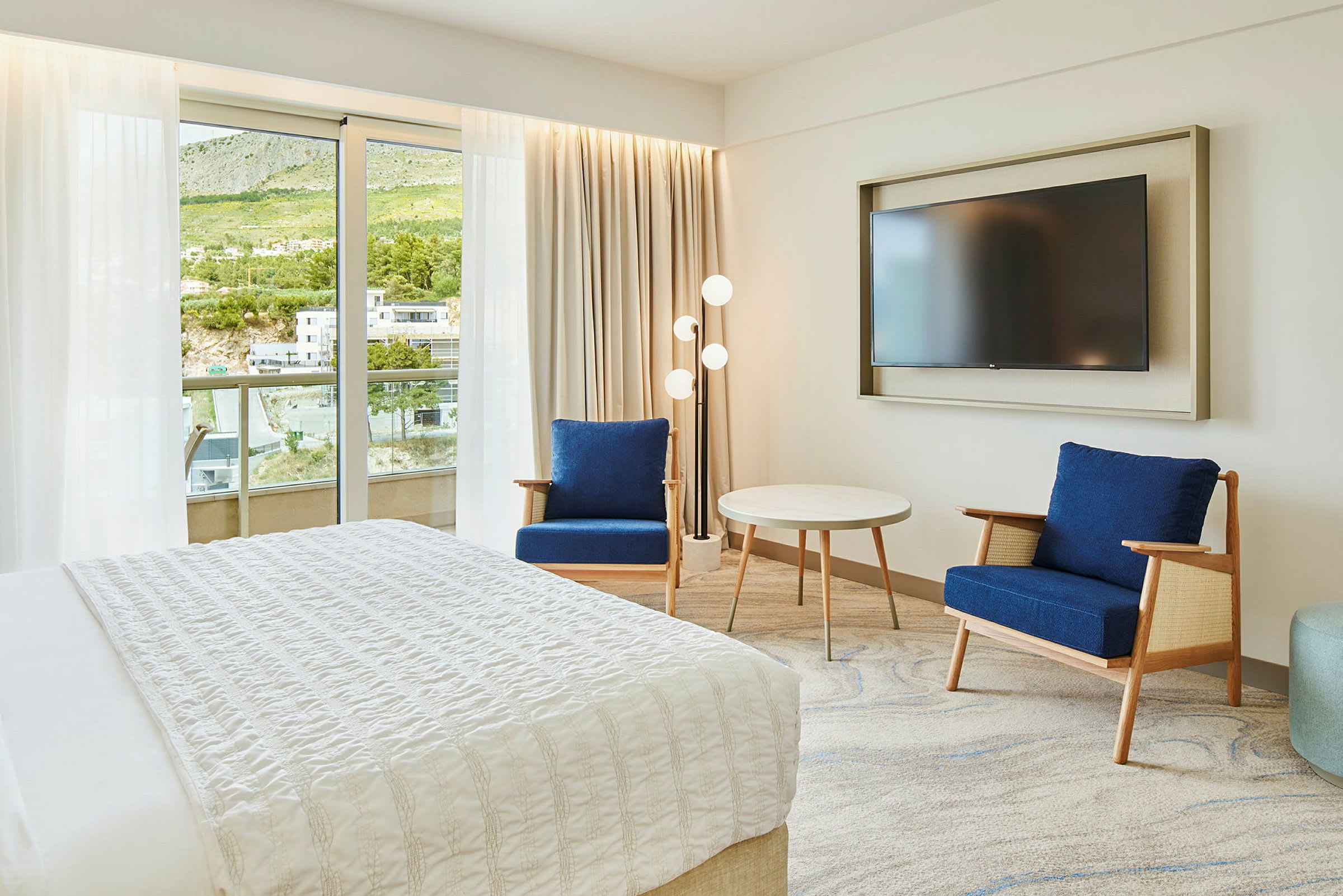 Hotellrom med seng, to blå stoler, et rundt bord og en veggmontert TV. Balkong og grønn utsikt ses gjennom glassdøren. (AI-generert tekst)
