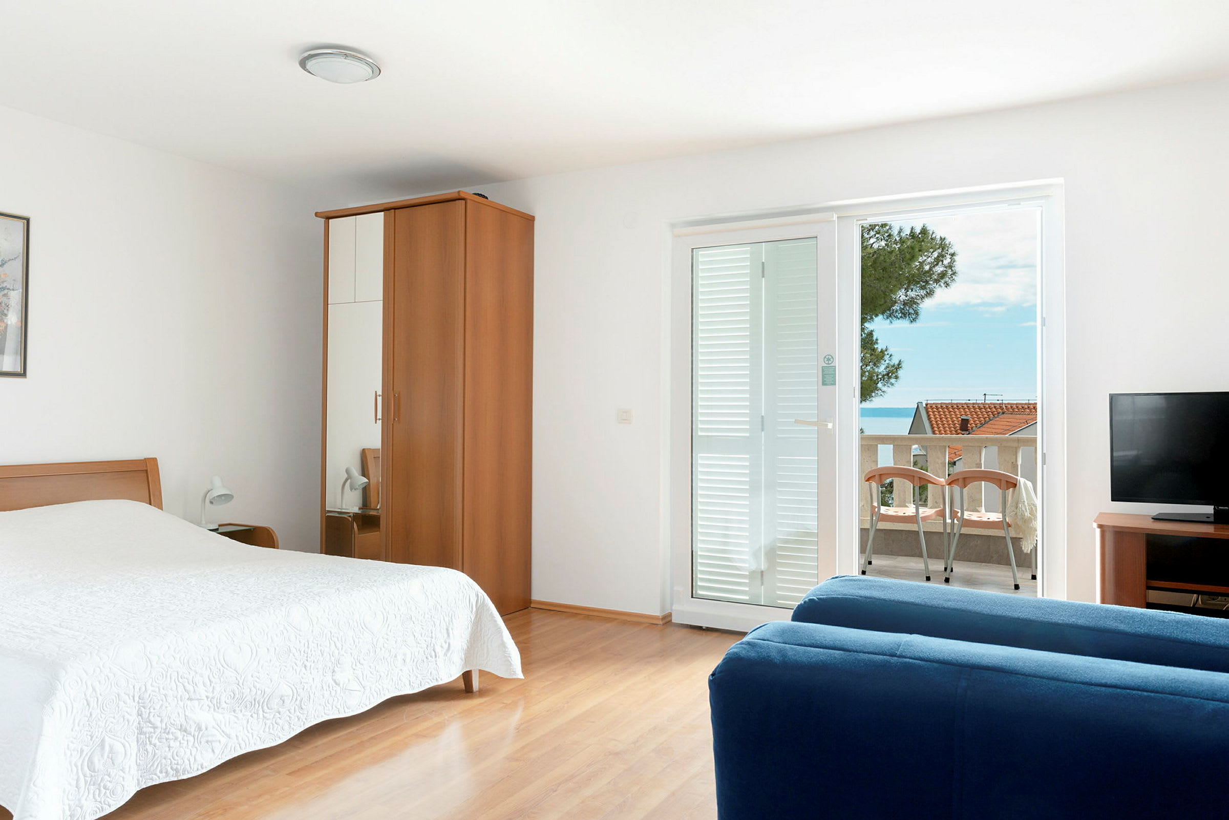 Et hotellrom med seng, garderobe, blå sofa, TV og balkong med utsikt. (AI-generert tekst)