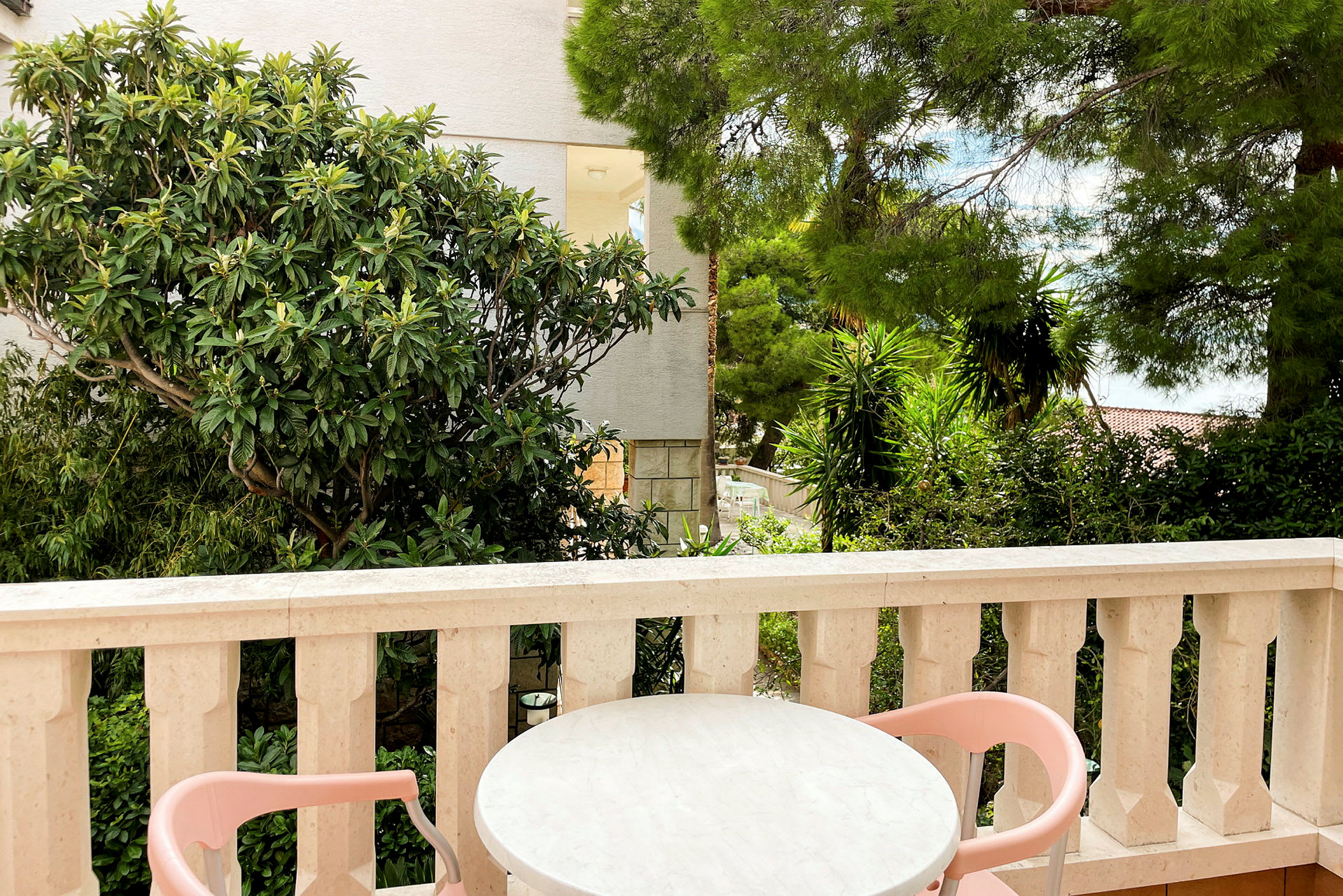 En terrasse med et bord og to rosa stoler, omgitt av grønne planter og trær. (AI-generert tekst)