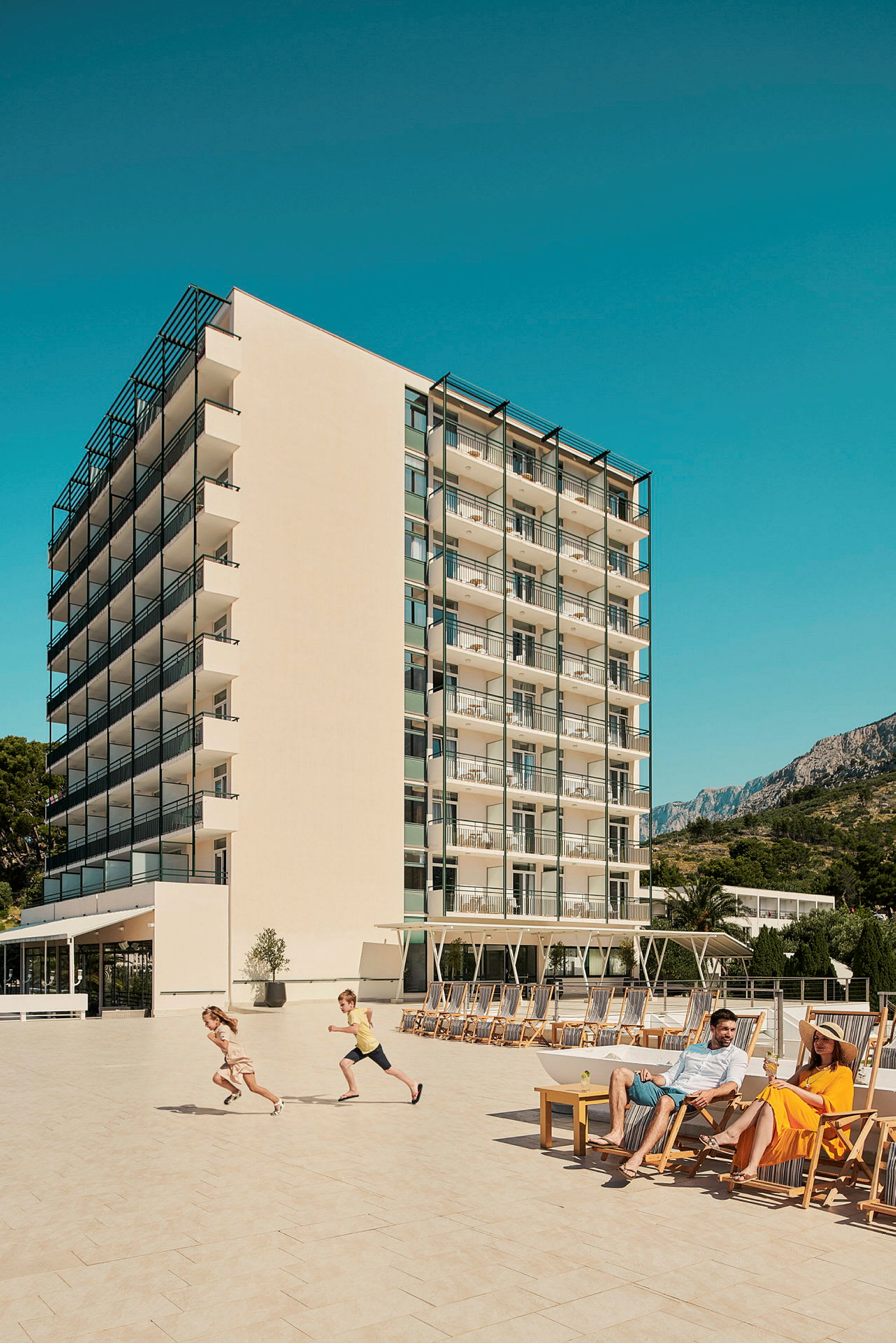 Bilde av et hotell med balkonger i flere etasjer, en stor terrasse med solsenger, og personer som nyter solen. (AI-generert tekst)