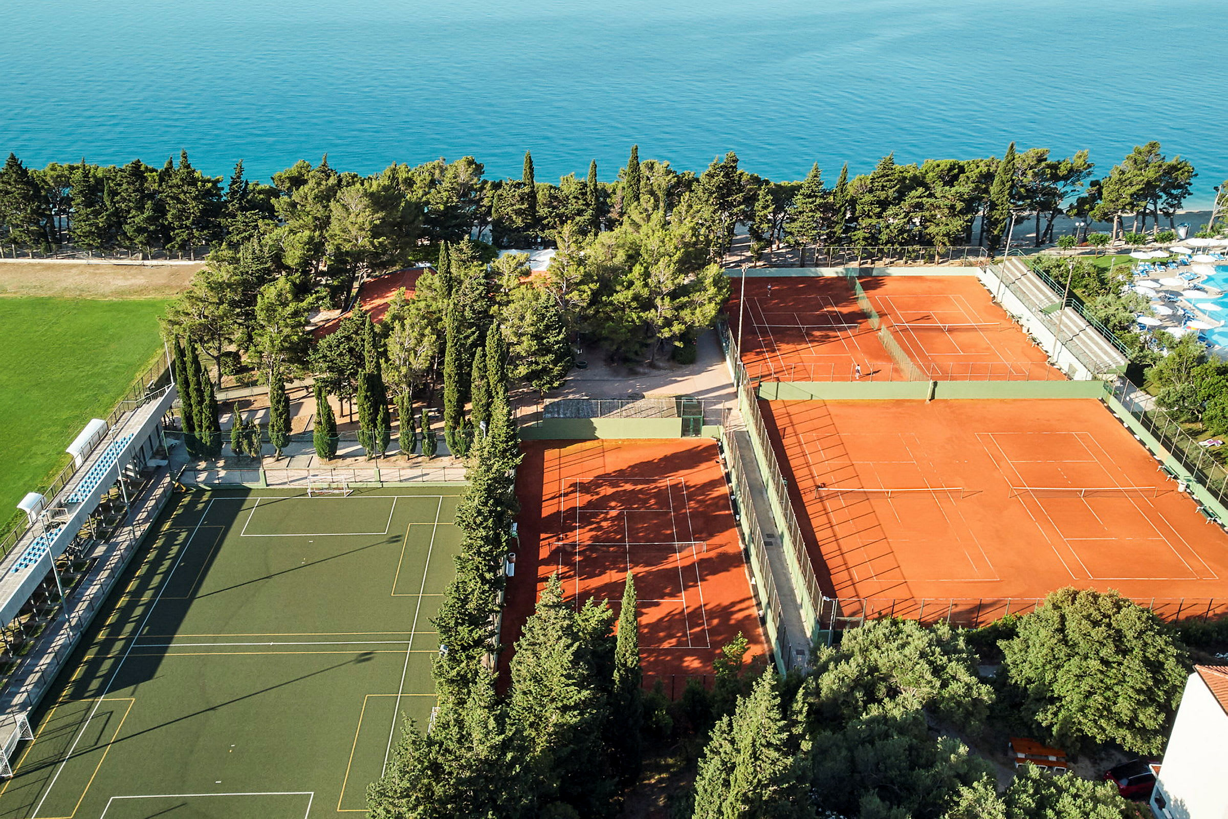 Luftfoto av idrettsanlegg med tennisbaner og fotballbane, omgitt av trær og hav. (AI-generert tekst)