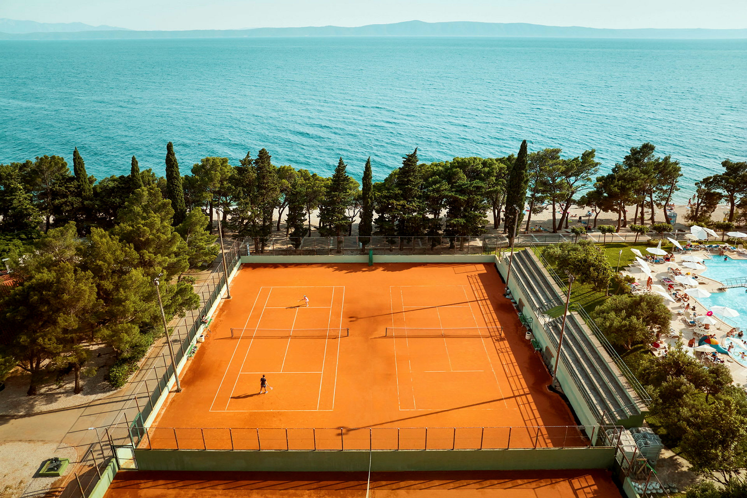 Tennisbane med spillere nær sjøen og et bassengområde. (AI-generert tekst)