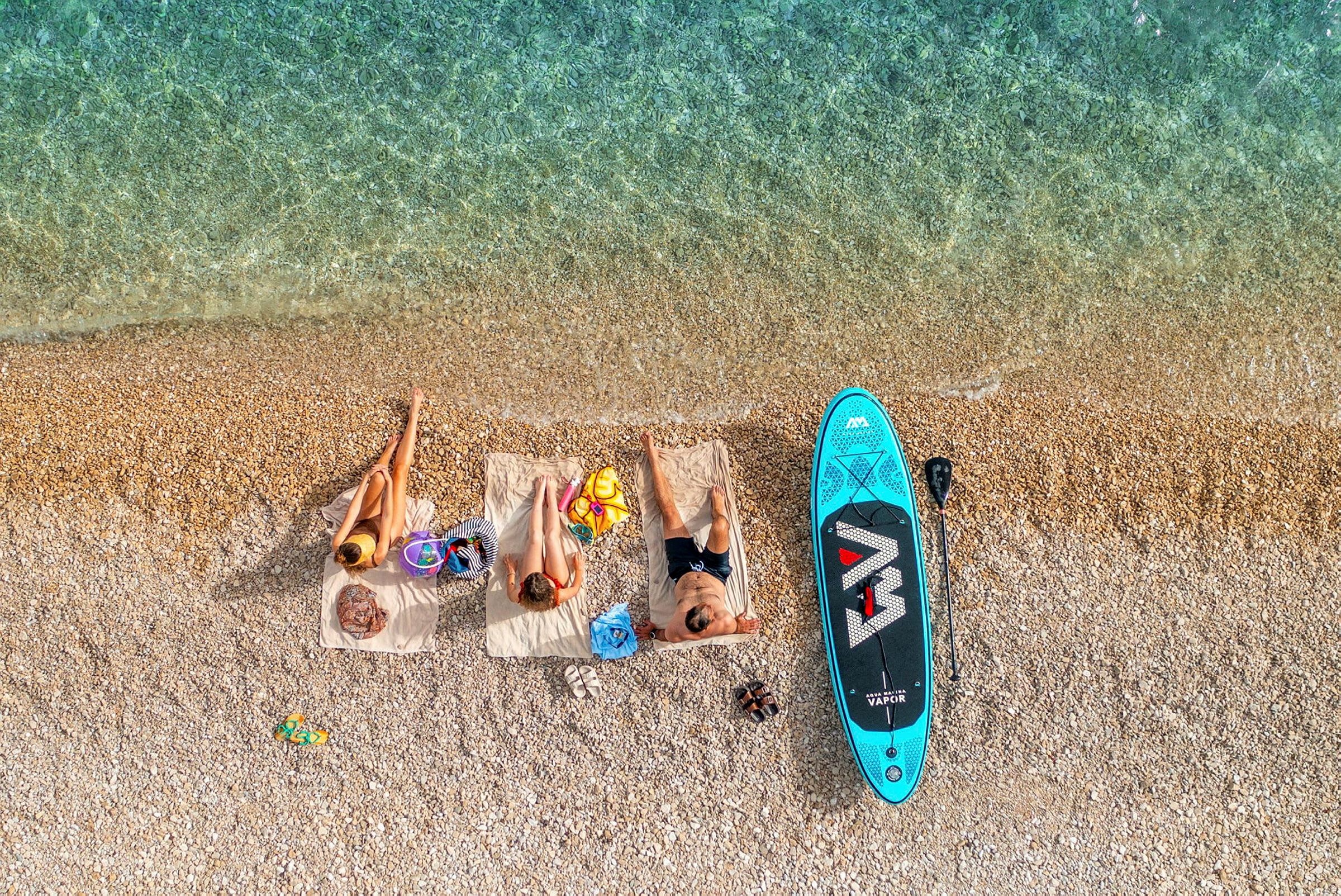Tre personer som ligger på håndklær på stranden ved siden av et paddleboard. (AI-generert tekst)