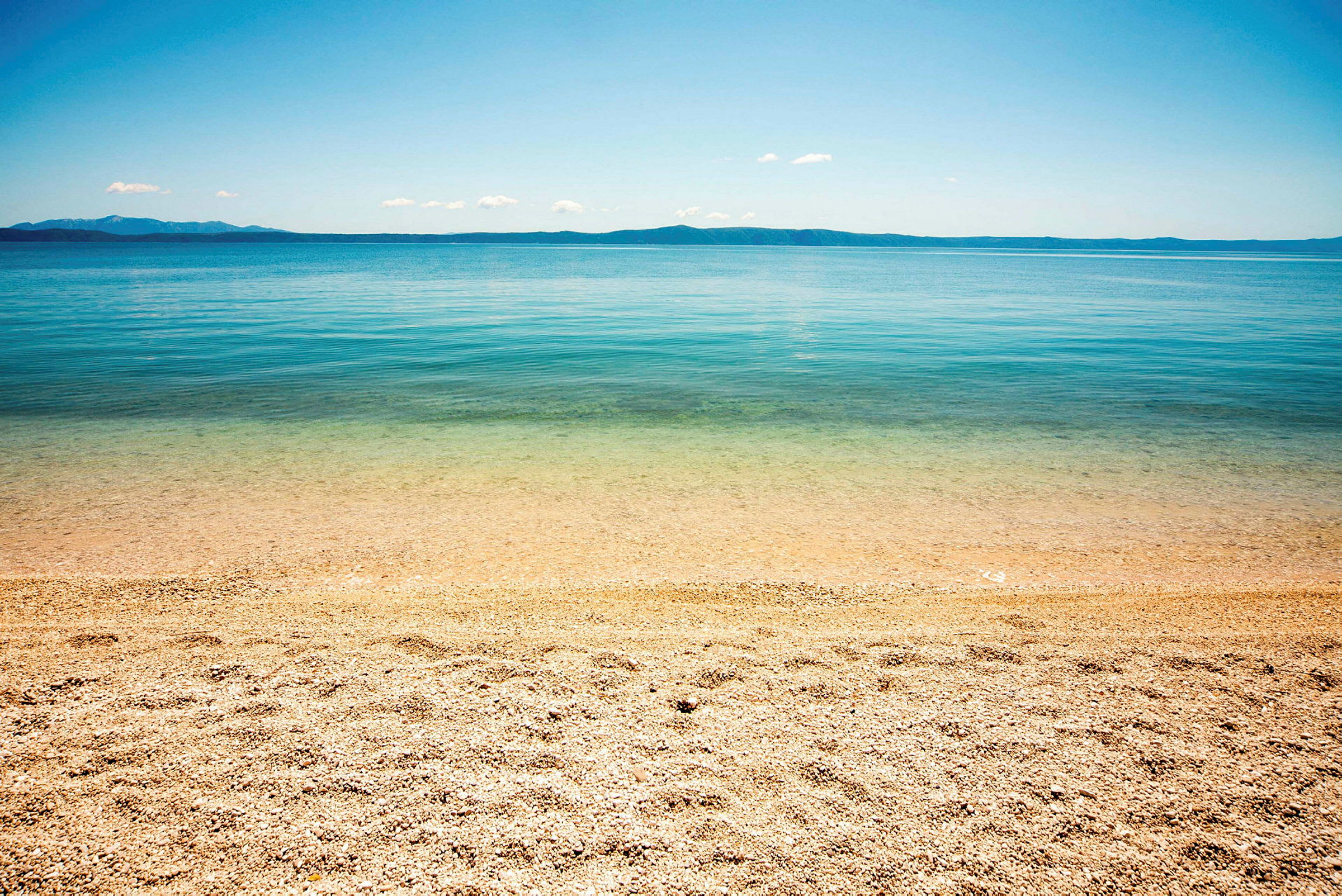 En strand med småstein ved klart vann og en fjern horisont. (AI-generert tekst)