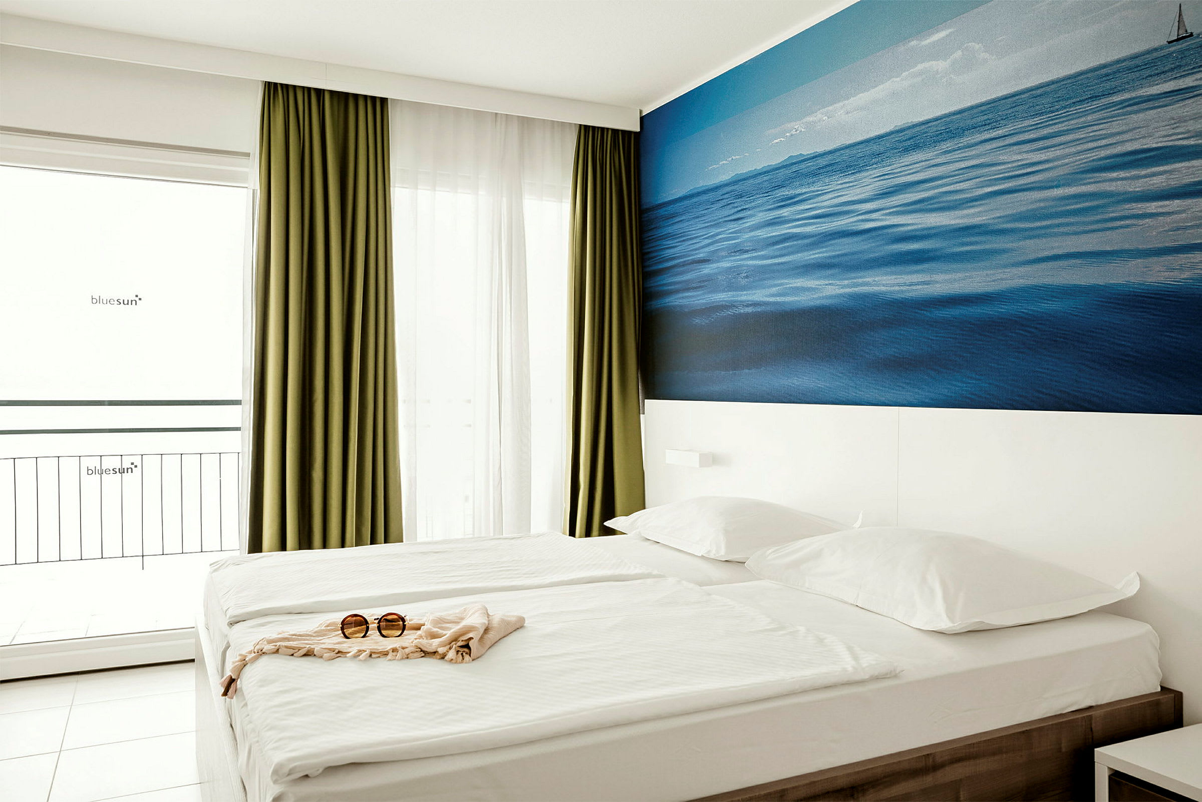 Et hotellrom med en dobbelseng, grønne gardiner, en hvit vegg med et blått bilde av havet og et par solbriller på sengen. (AI-generert tekst)