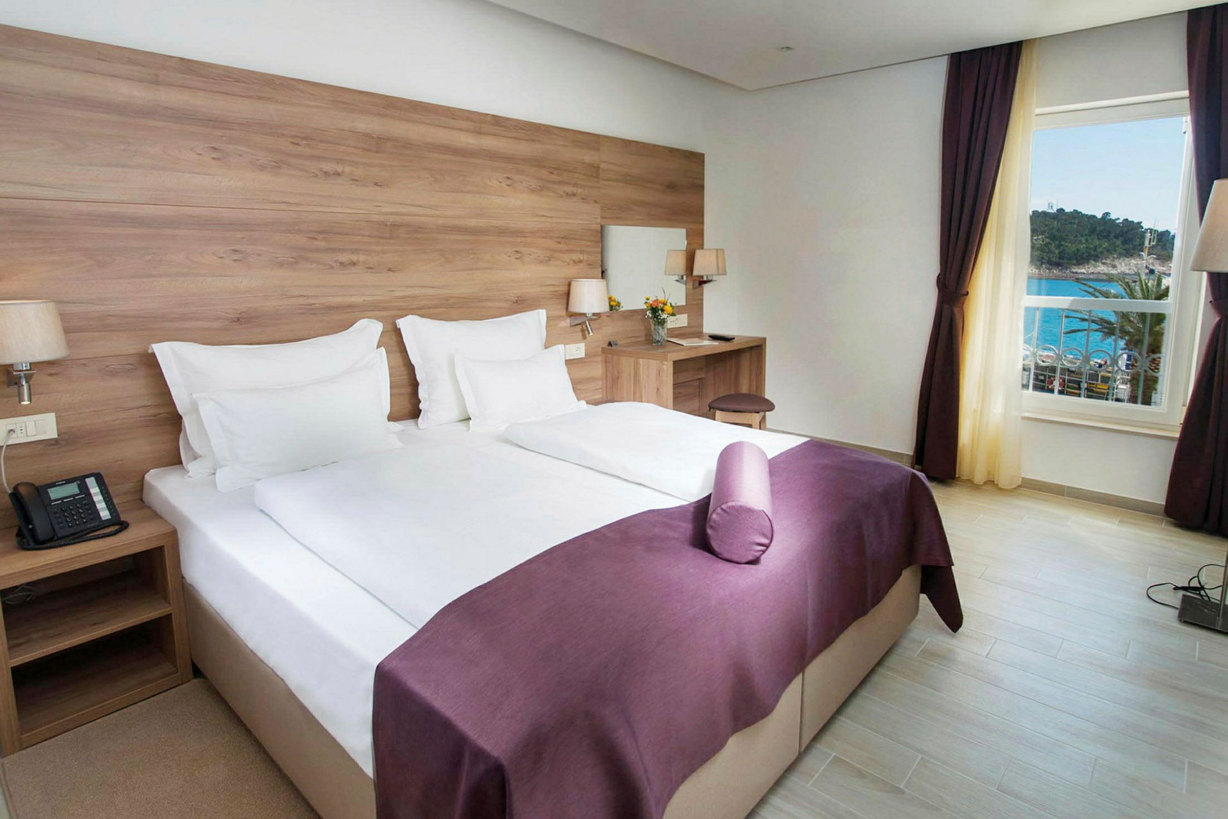 Et hotellrom med en dobbeltseng, hvite laken og lilla sengeteppe. Det er et skrivebord med en stol og et speil. Vinduet viser utsikt mot havet. (AI-generert tekst)
