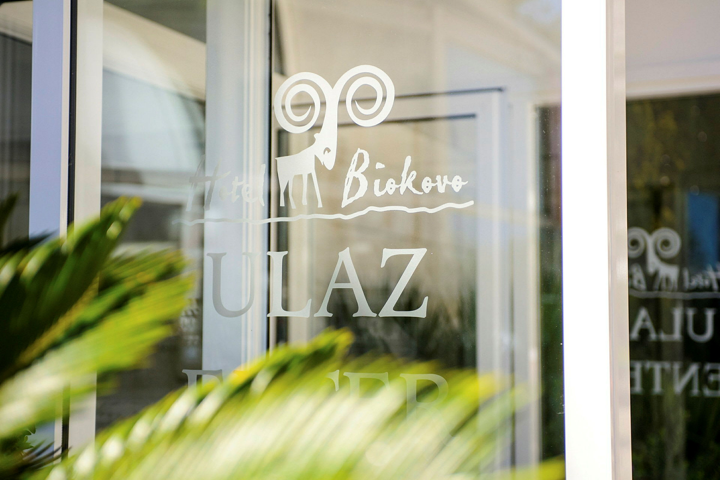 Tekst på en glassdør med skriften "Hotel Biokovo" og "ULAZ," med en tegnet logo av en steinbukk. (AI-generert tekst)
