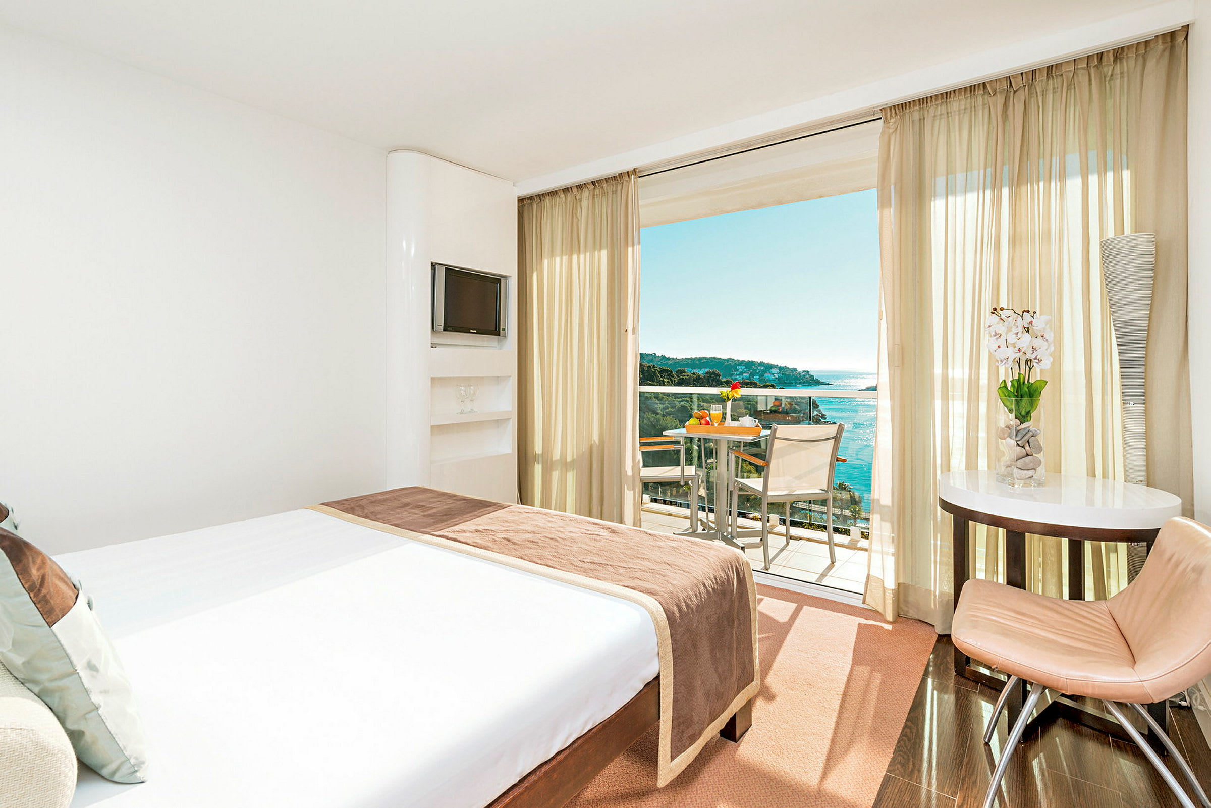Hotellrom med en stor seng vendt mot en balkong med utsikt over havet. (AI-generert tekst)