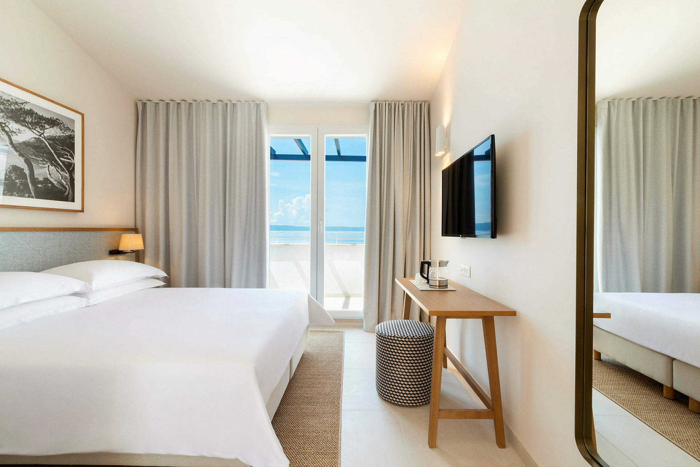 Et hotellrom med en stor seng, et skrivebord og en TV på veggen. Store vinduer med utsikt mot havet. (AI-generert tekst)