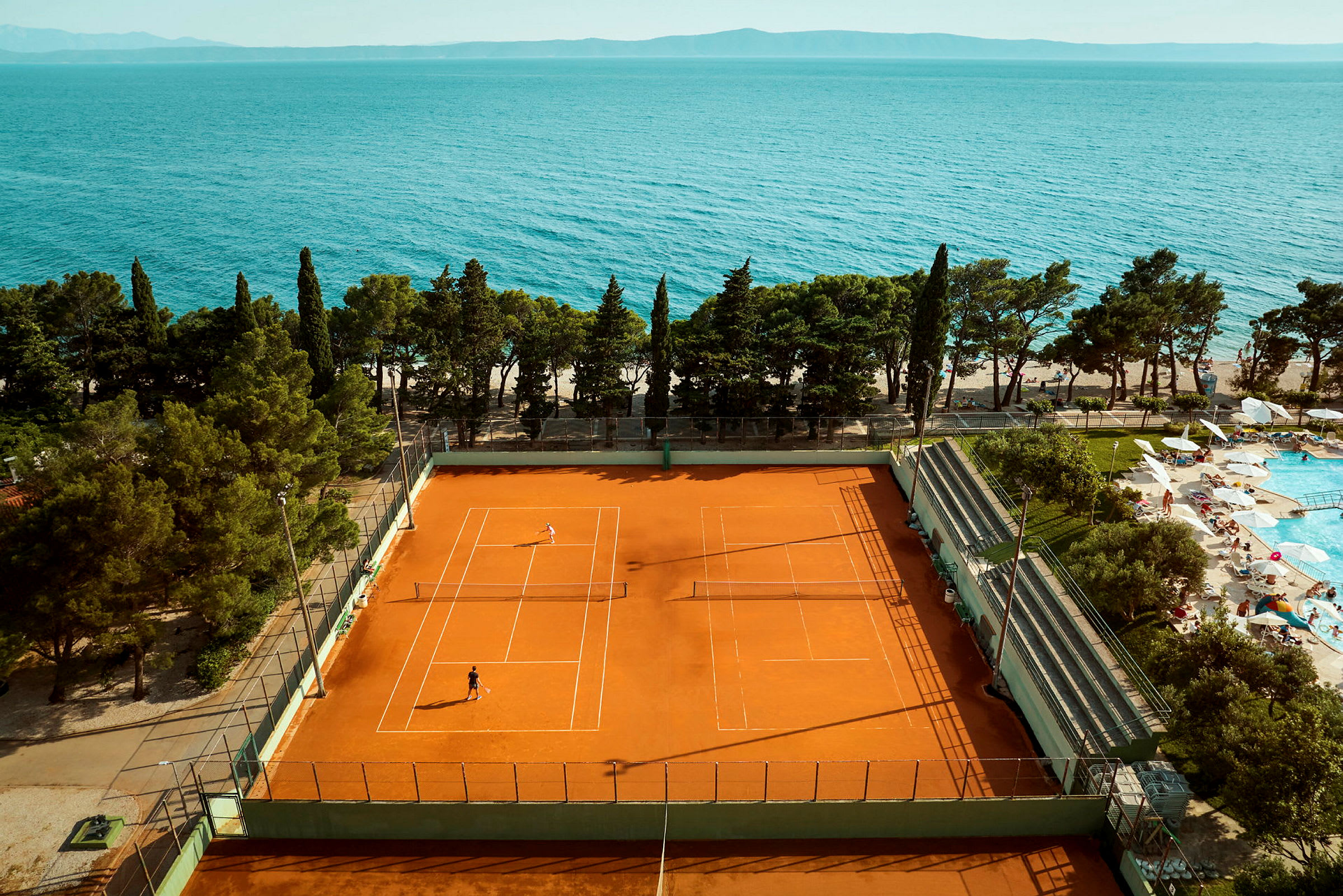 En tennisbane med spillere og et bassengområde med solsenger ved siden av, samt hav i bakgrunnen. (AI-generert tekst)