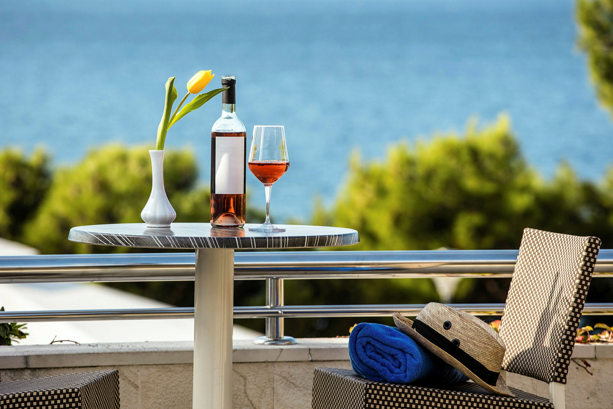 Et bord med en flaske og et glass rosévin samt en gul tulipan i vase, med utsikt over havet i bakgrunnen. (AI-generert tekst)