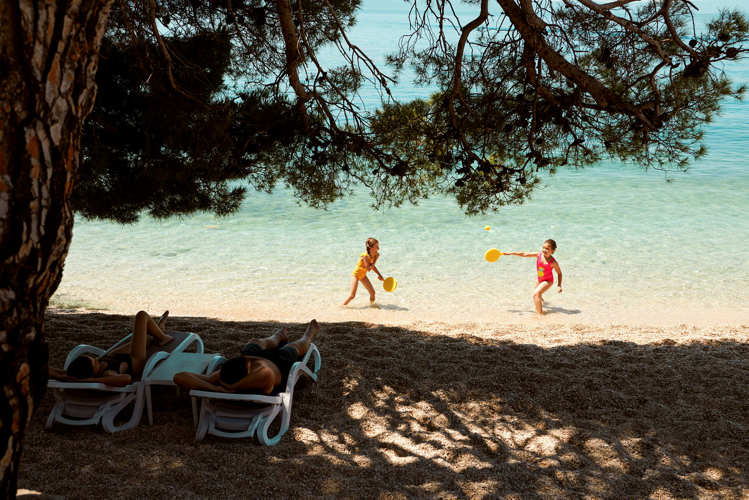 To barn leker med racketer i vannkanten på en strand. Flere personer ligger på solsenger under skyggefulle trær. (AI-generert tekst)