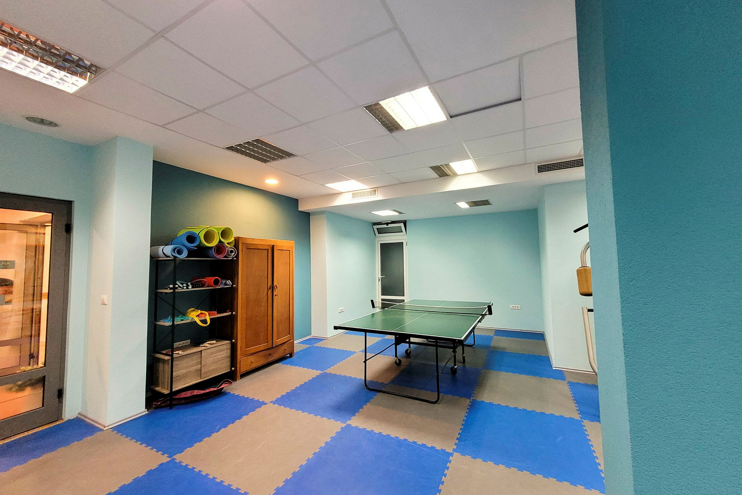 Et bilde av et rom med bordtennisbord og hylle med yogamatter. (AI-generert tekst)