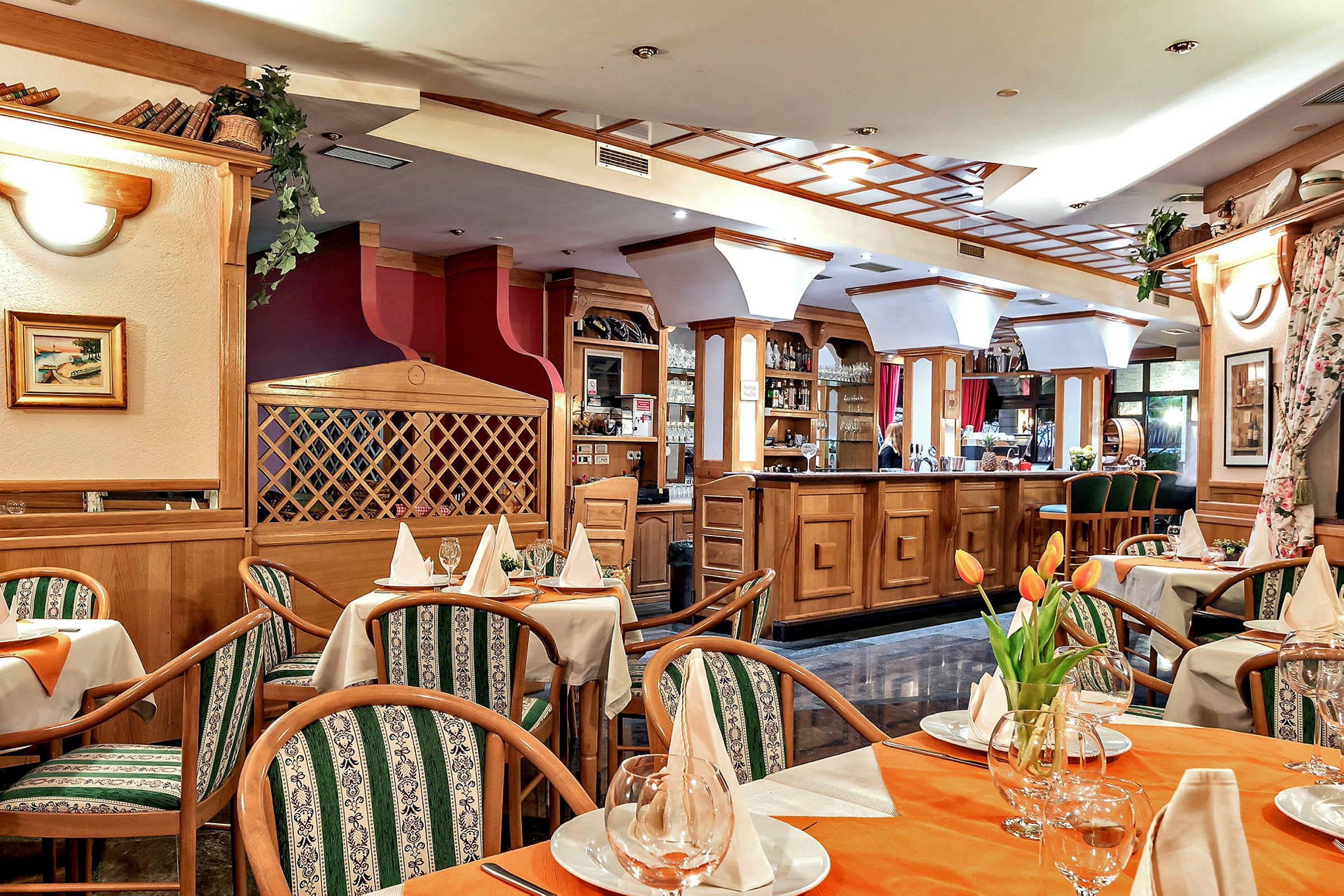 En innredet restaurant med bord dekket med hvite servietter og glass. Stoler med grønne mønstrede puter omgir bordene, og en bardisk i tre er synlig i bakgrunnen. (AI-generert tekst)
