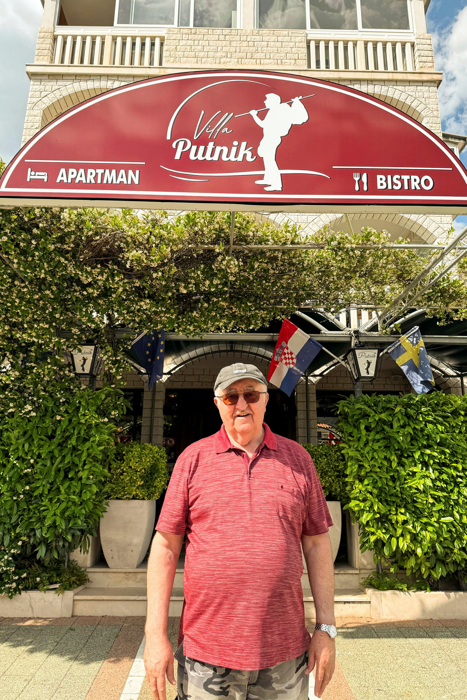 Et bilde av en person som står foran en bygning med et skilt som sier "Villa Putnik", omgitt av grønne planter. (AI-generert tekst)
