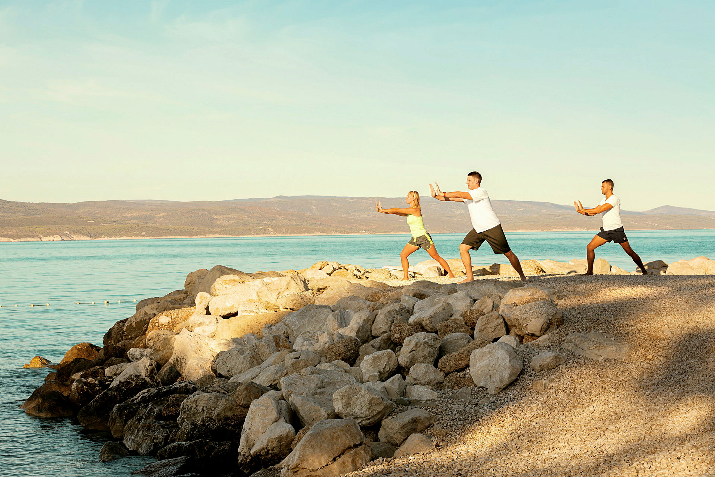 Tre personer utfører tai chi ved stranden ved sjøen. (AI-generert tekst)