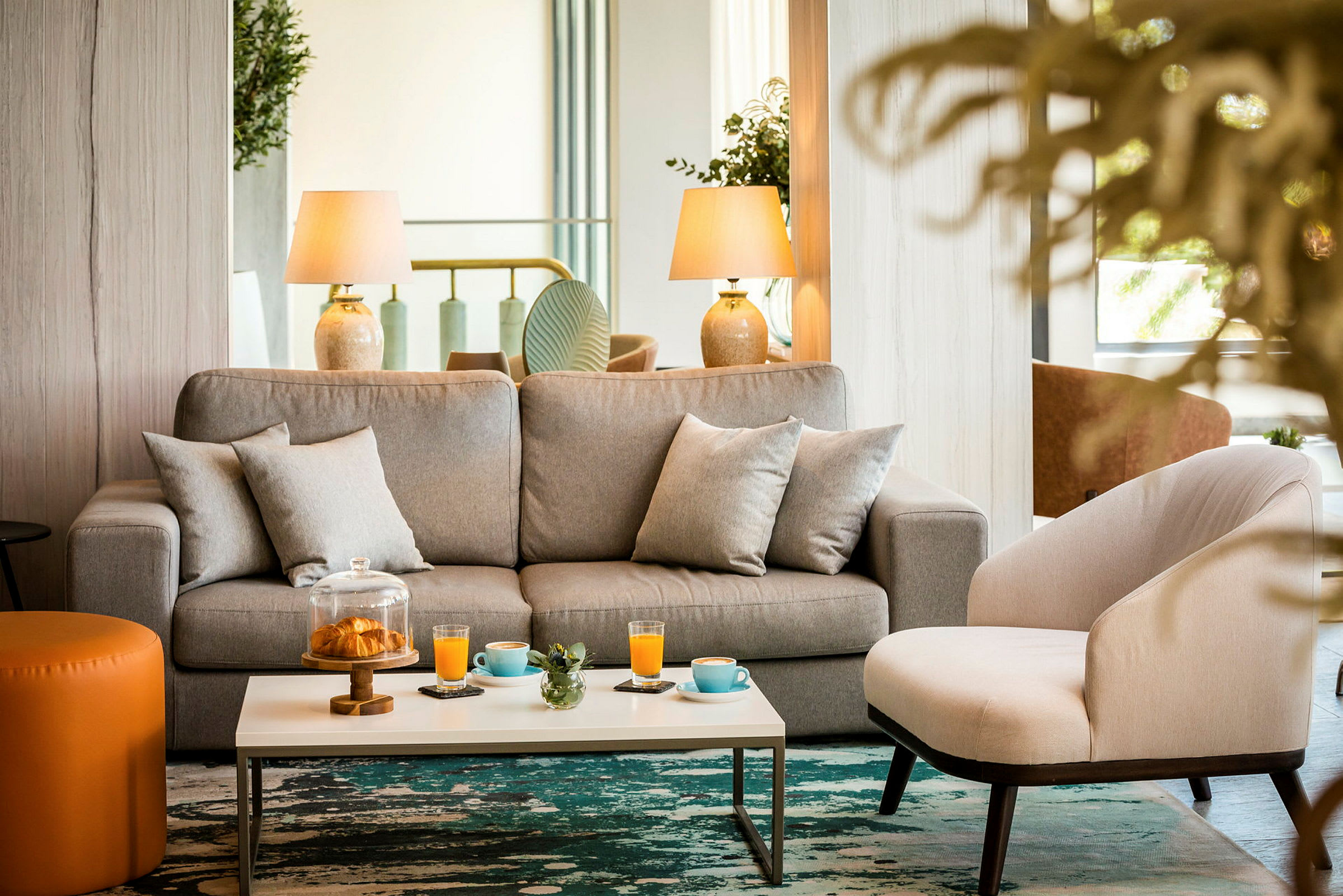 En stue med en grå sofa og en beige lenestol foran et bord med drikker og en glassbeholder med bakverk. (AI-generert tekst)