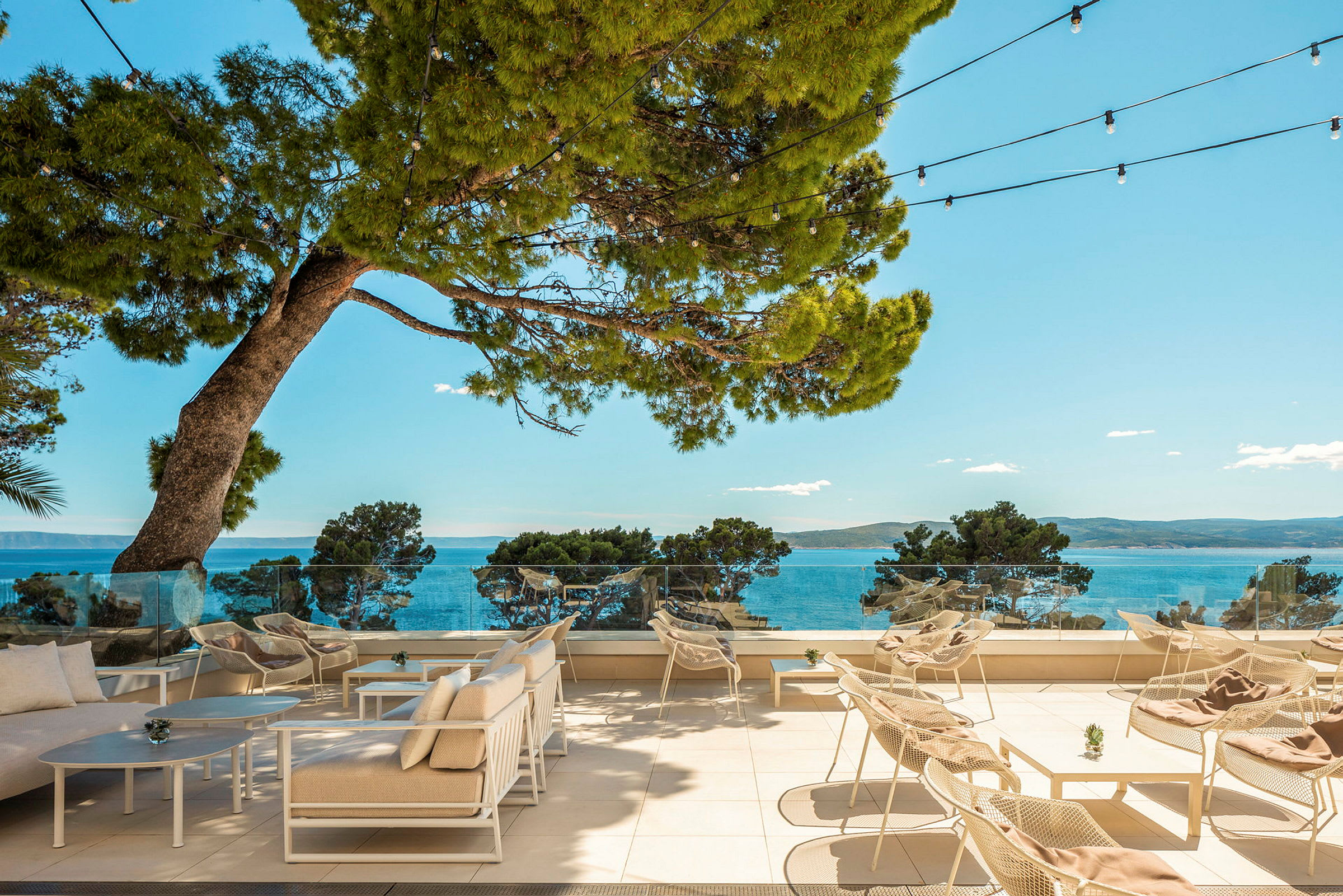 Utendørs terrasse med stoler og bord, med utsikt mot havet og omkringliggende trær. (AI-generert tekst)