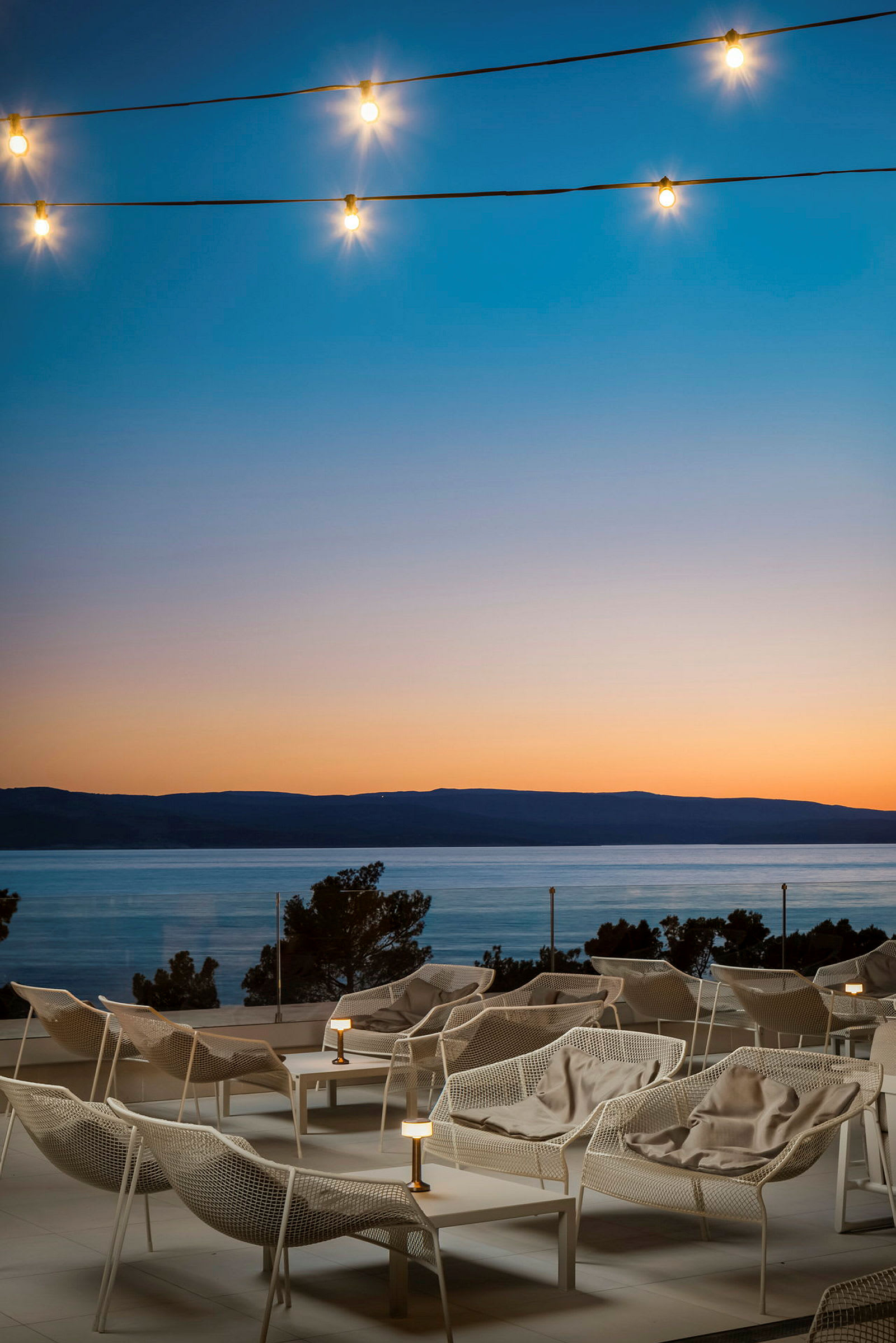 Hvite lounge-stoler på en terrasse med utsikt over havet ved solnedgang, lys til dekorasjon. (AI-generert tekst)