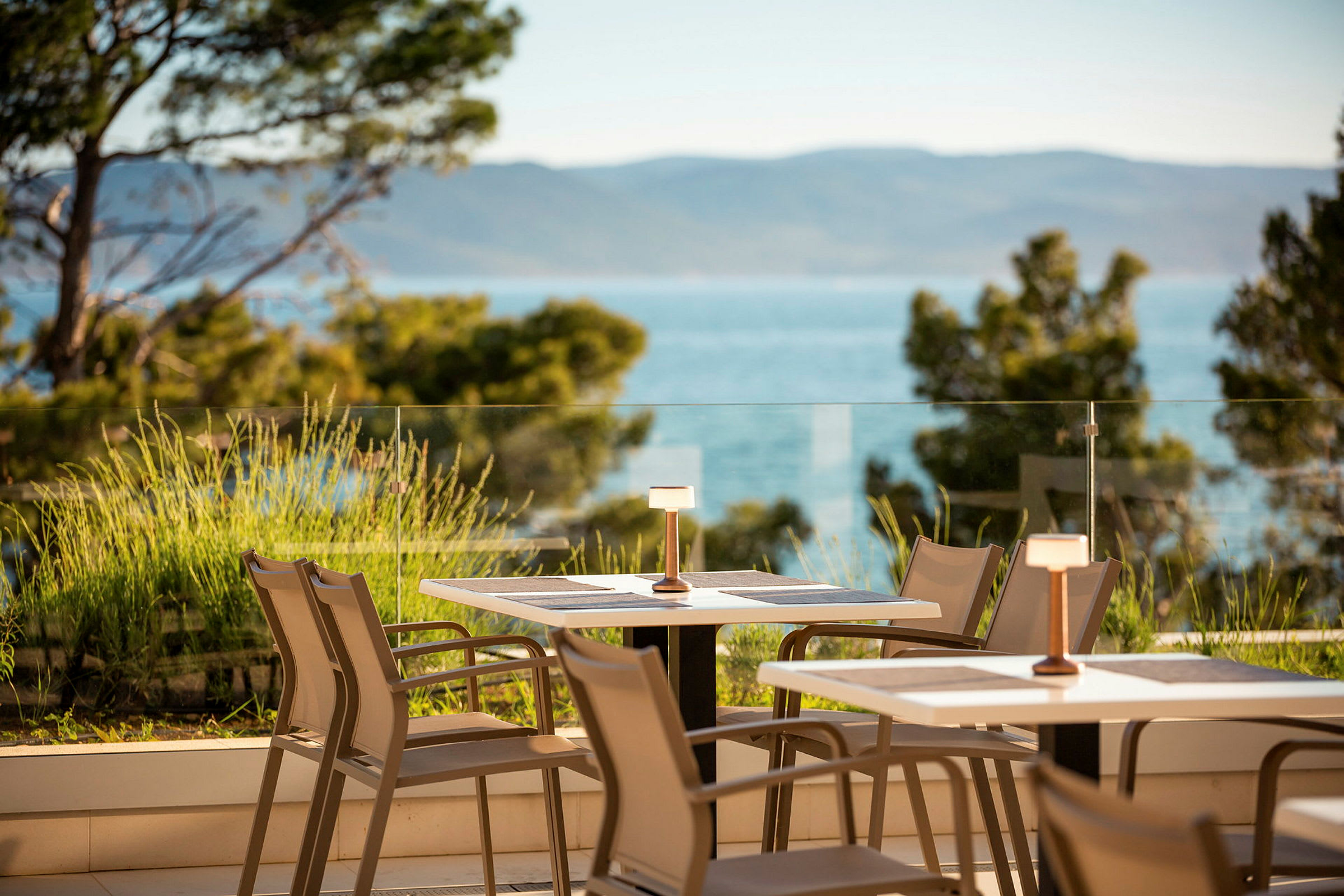 Utsikt over en terrasse med bord og stoler, omgitt av grøntområder og med havet i bakgrunnen. (AI-generert tekst)