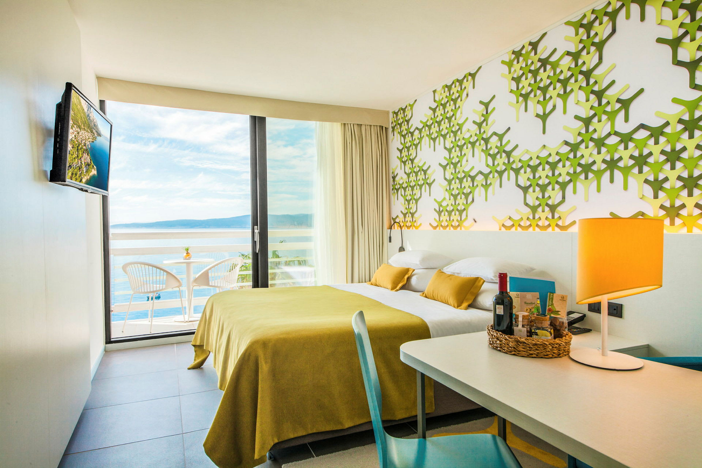 Hotellrom med moderne innredning, gul sengeteppe, grønn mønstret vegg og utsikt over havet fra balkongen. (AI-generert tekst)