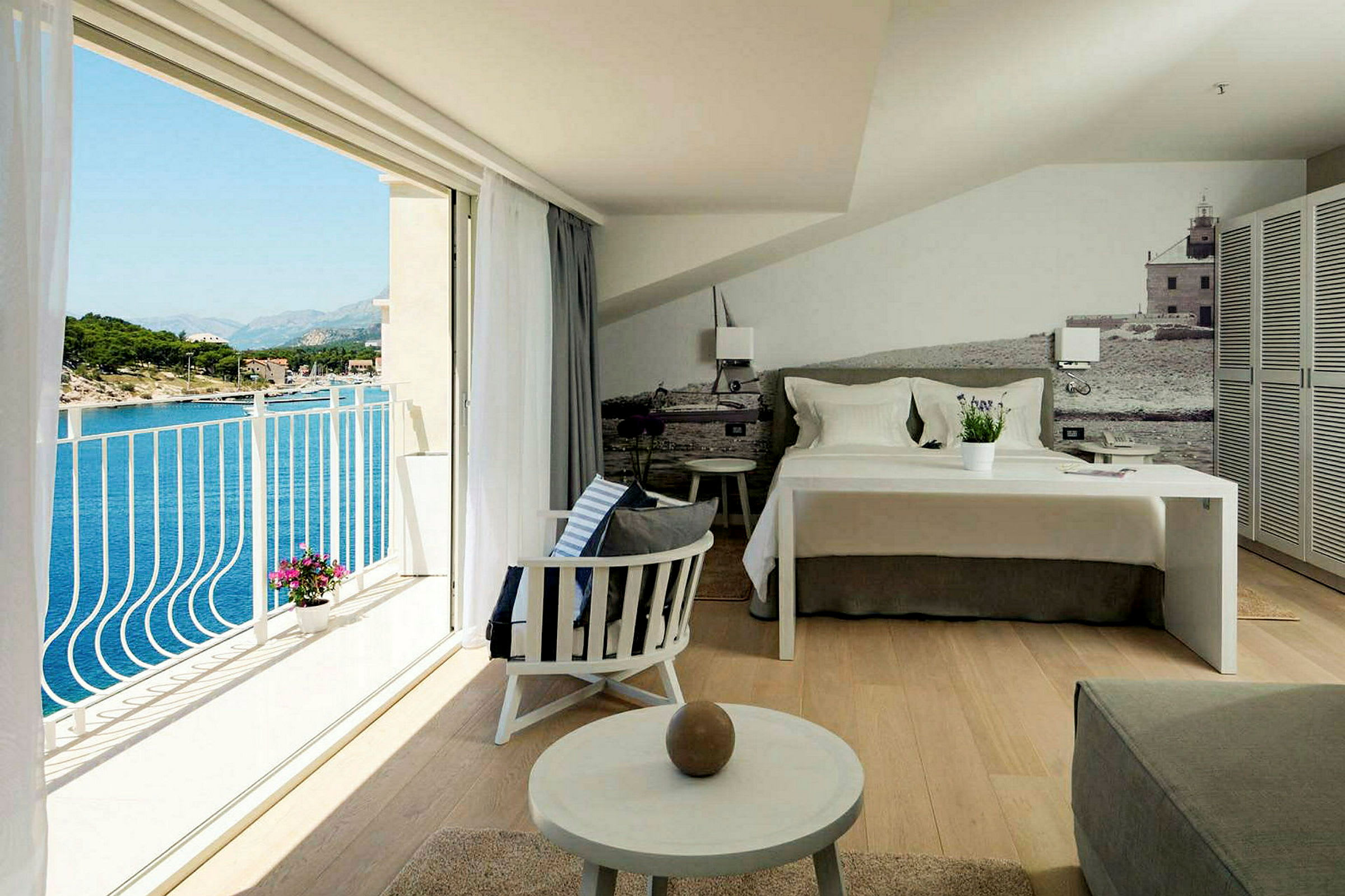 Et hotellrom med en oppredd seng, sitteområde og en balkong med utsikt over havet. (AI-generert tekst)