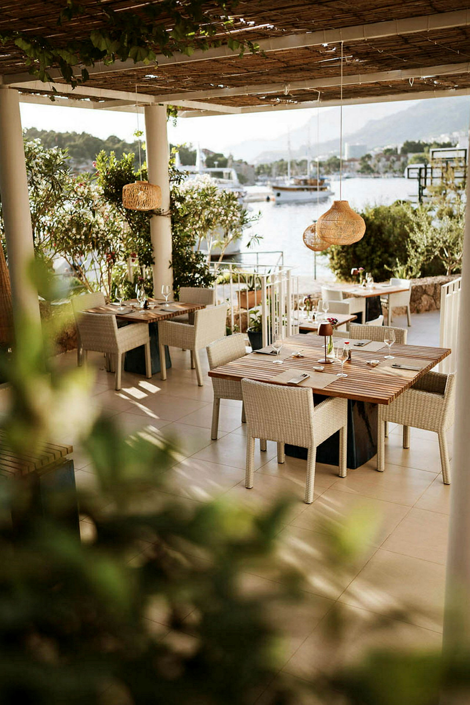 En utendørs restaurant med bord og stoler under tak, nær vann med båter i bakgrunnen. (AI-generert tekst)