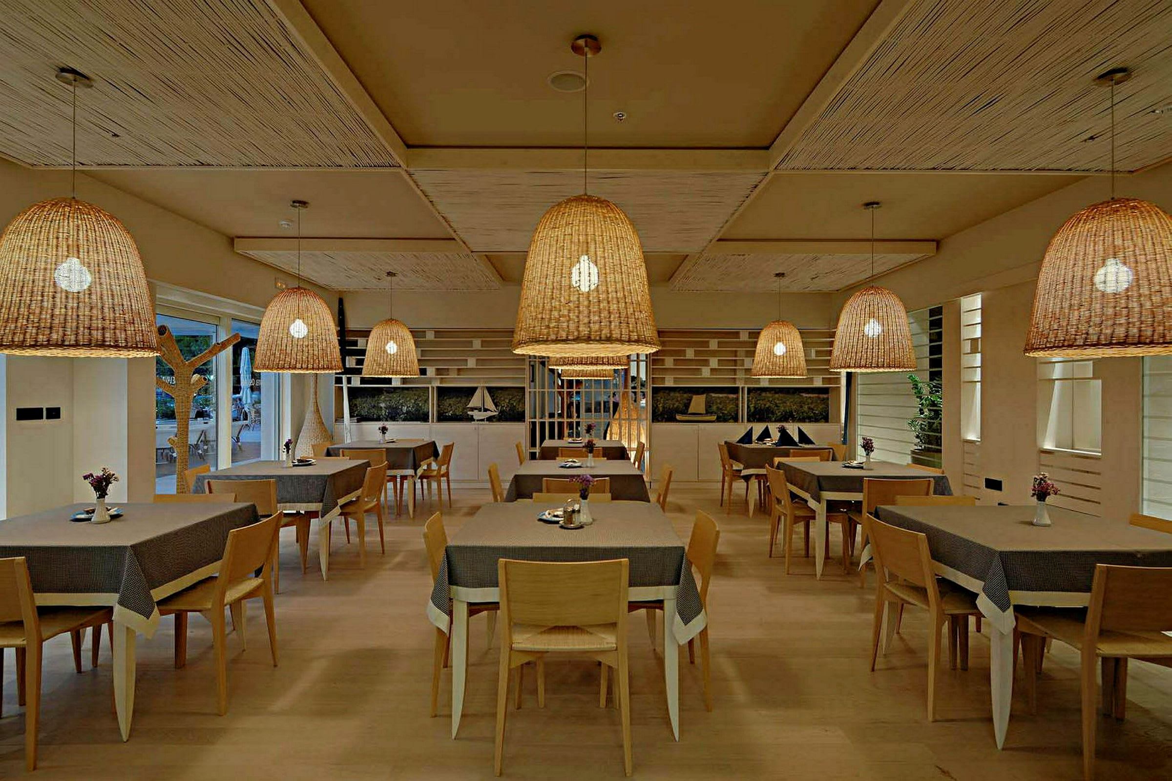 En restaurant med flere bord og stoler samt store flettede taklamper. (AI-generert tekst)