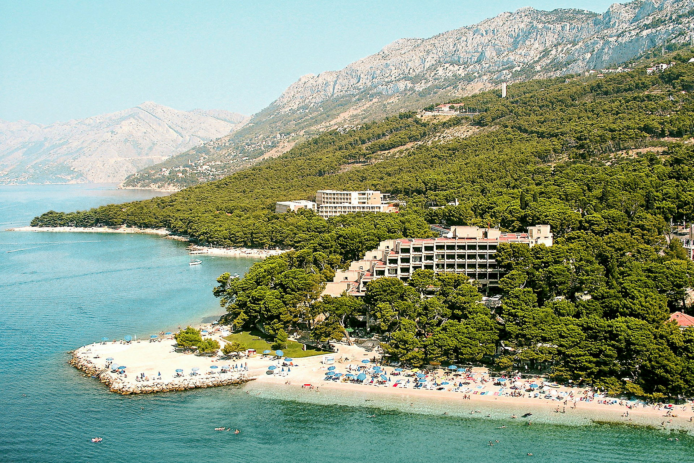 Strand, hotell og vegetasjon ved en kyst med fjell i bakgrunnen. (AI-generert tekst)