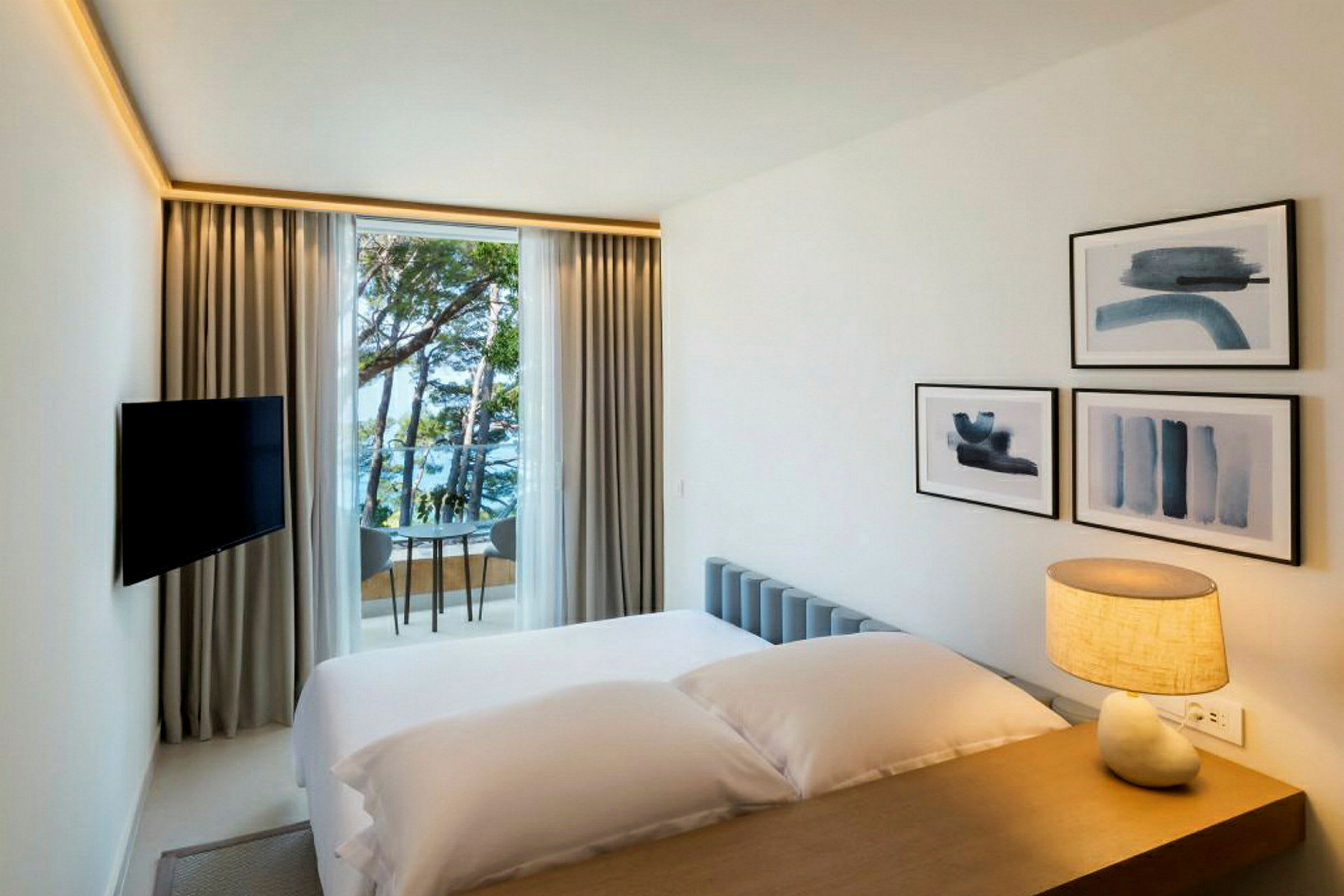 Et hotellrom med en seng, en lampe på bordet, veggdekorasjoner og et vindu som vender mot havet og trær. (AI-generert tekst)
